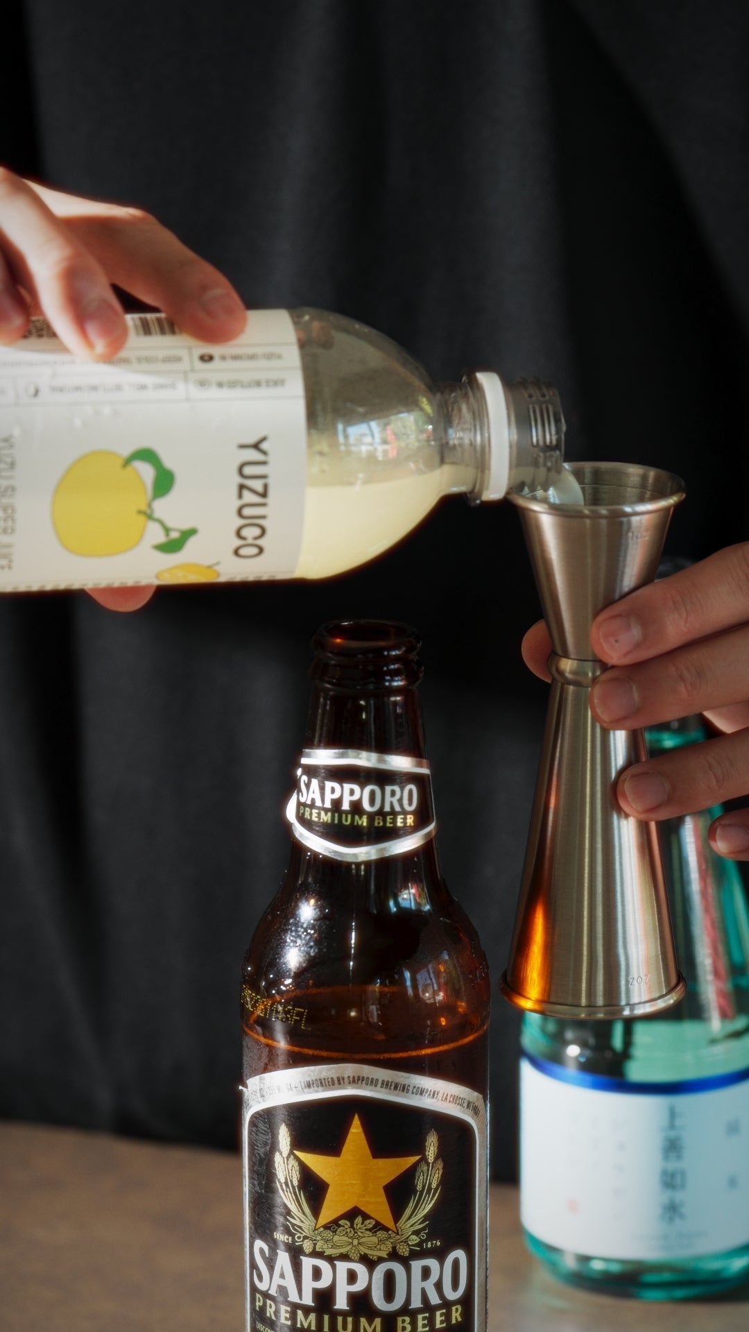 Hands pouring YUZUCO yuzu juice into a metal jigger over a Sapporo beer bottle on a countertop