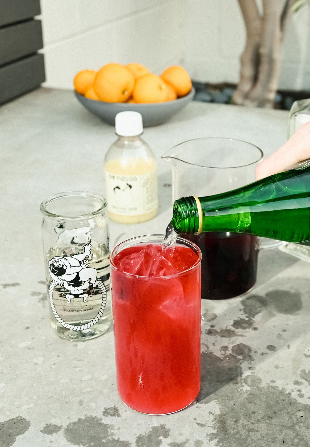 Hand pouring sparkling water into an iced glass with bright red yuzu cocktail, Yuzuco bottle and bowl of oranges on counter