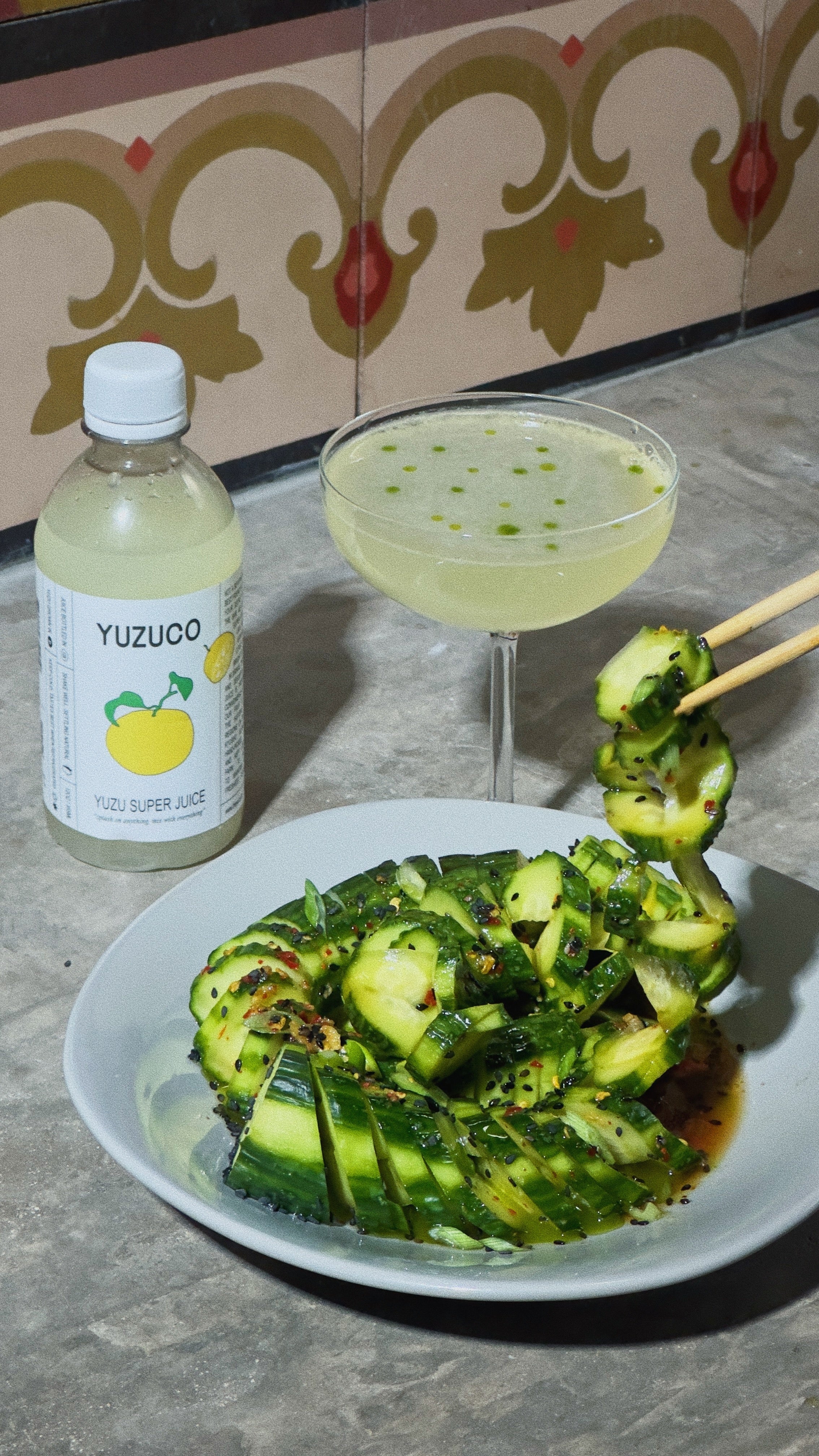 Yuzu cucumber salad tossed with sesame, chili flakes, and scallions next to a yuzu gimlet cocktail and a YUZUCO bottle on a tiled counter