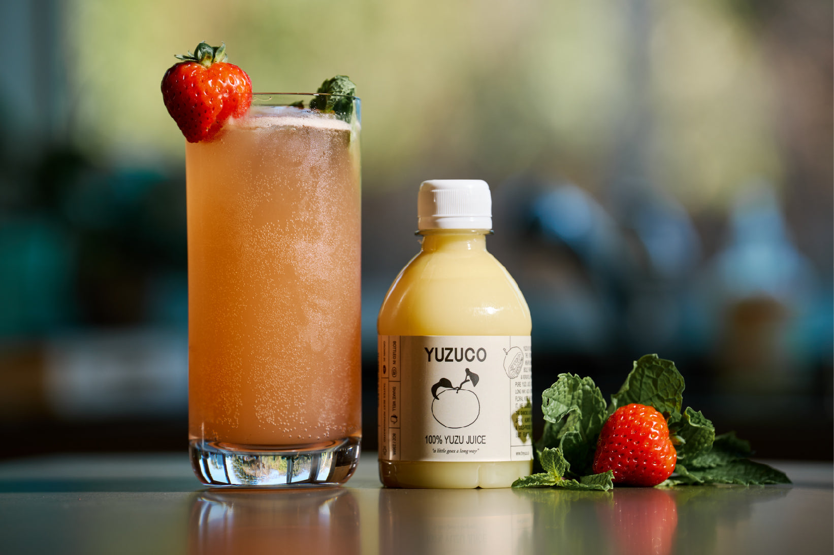 Pink yuzu cocktail in a tall glass garnished with a strawberry, beside a bottle of YUZUCO 100% yuzu juice and mint on a countertop