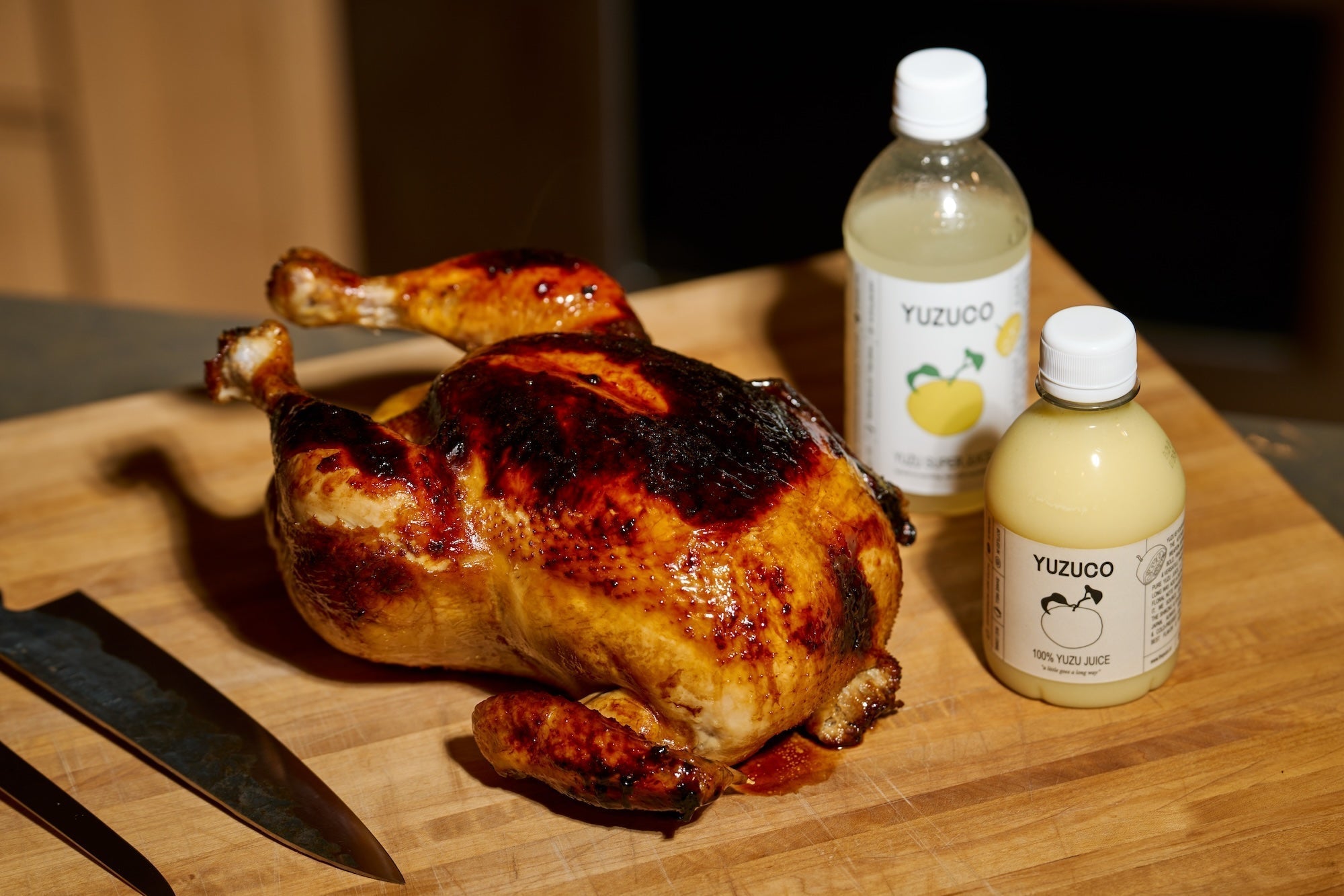 Roasted whole chicken glazed with yuzu honey on a wooden cutting board with two YUZUCO yuzu juice bottles and carving knife