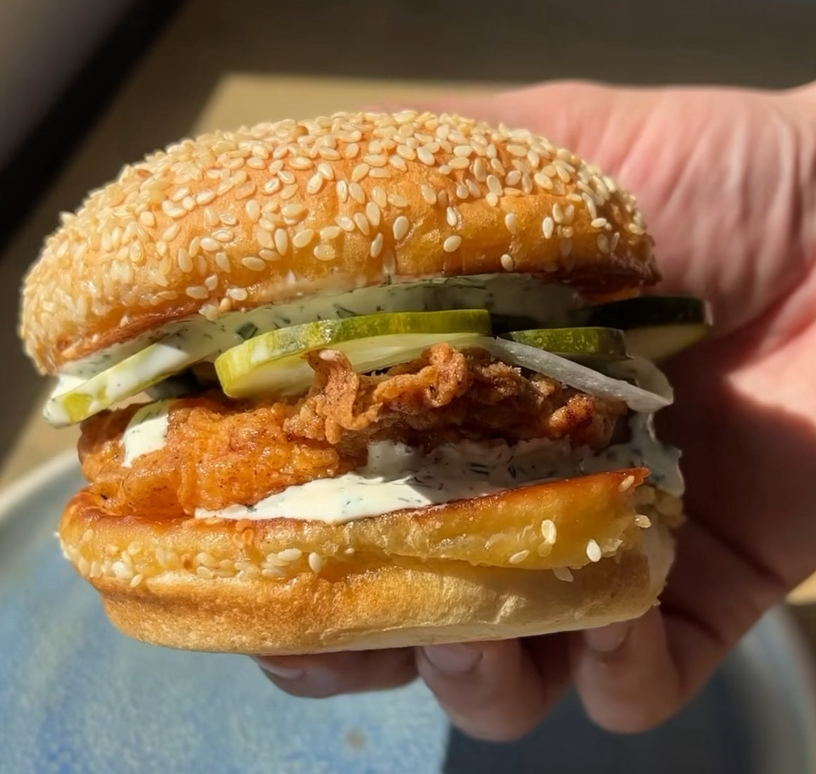 Close-up of sesame seed chicken sandwich with crispy fried chicken, yuzu ranch dressing, pickles and onions held in hand