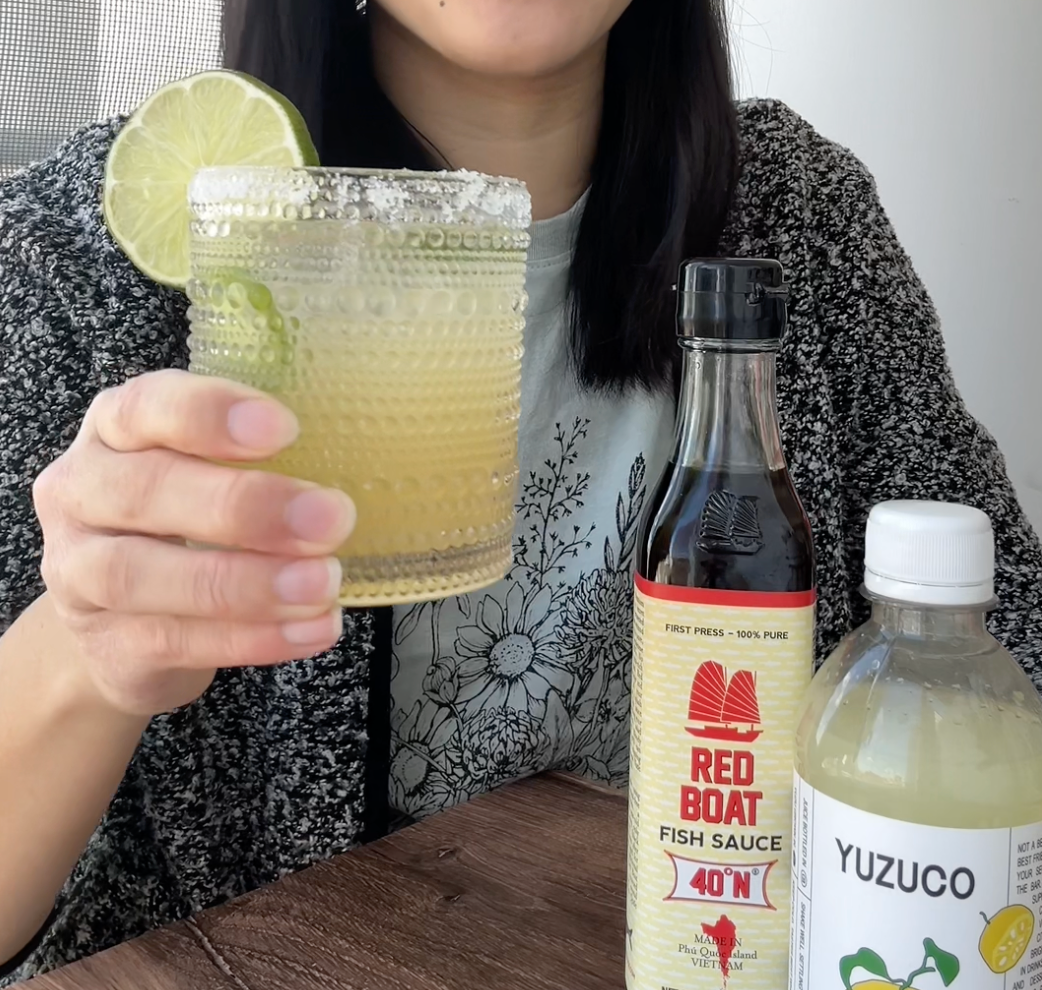 Person holding a salted-rim yuzu margarita garnished with a lime, beside a bottle of Red Boat fish sauce and a YUZUCO yuzu juice bottle on a wooden table