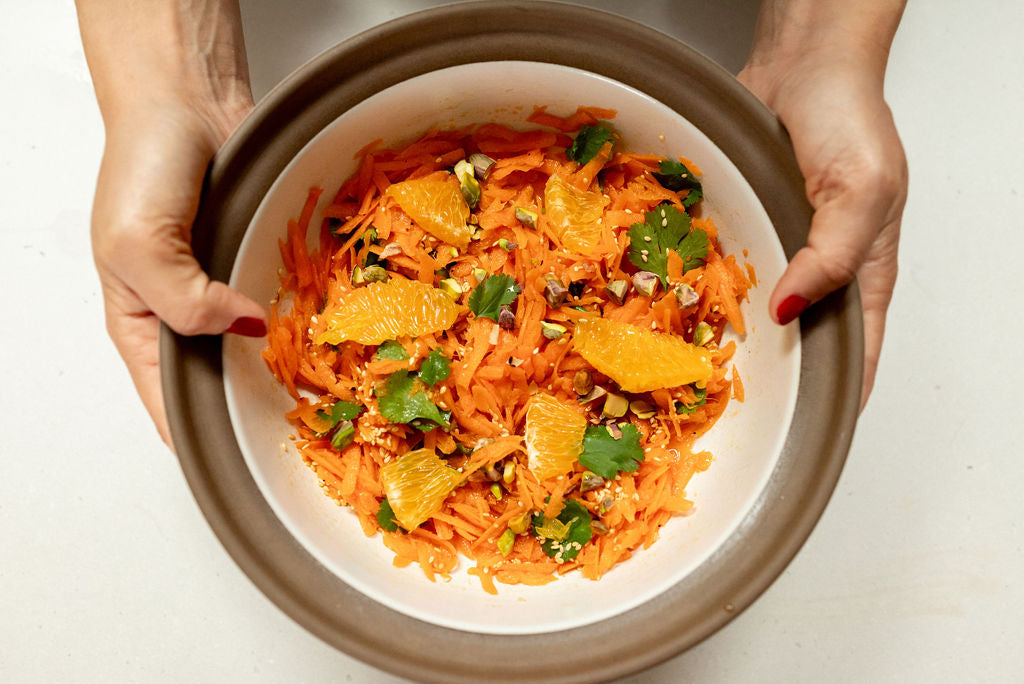 Top-down view of carrot salad with yuzu dressing showing shredded carrots, orange segments, cilantro and chopped pistachios in a white bowl held over a light surface