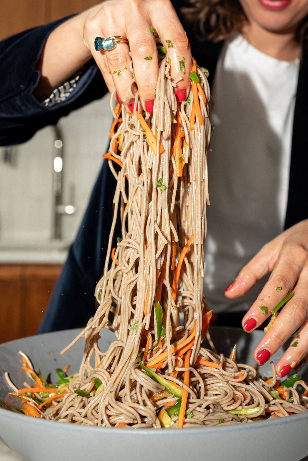 Hands with red nail polish mixing cold soba salad with yuzu-ginger dressing, shredded carrot and cucumber in a gray bowl