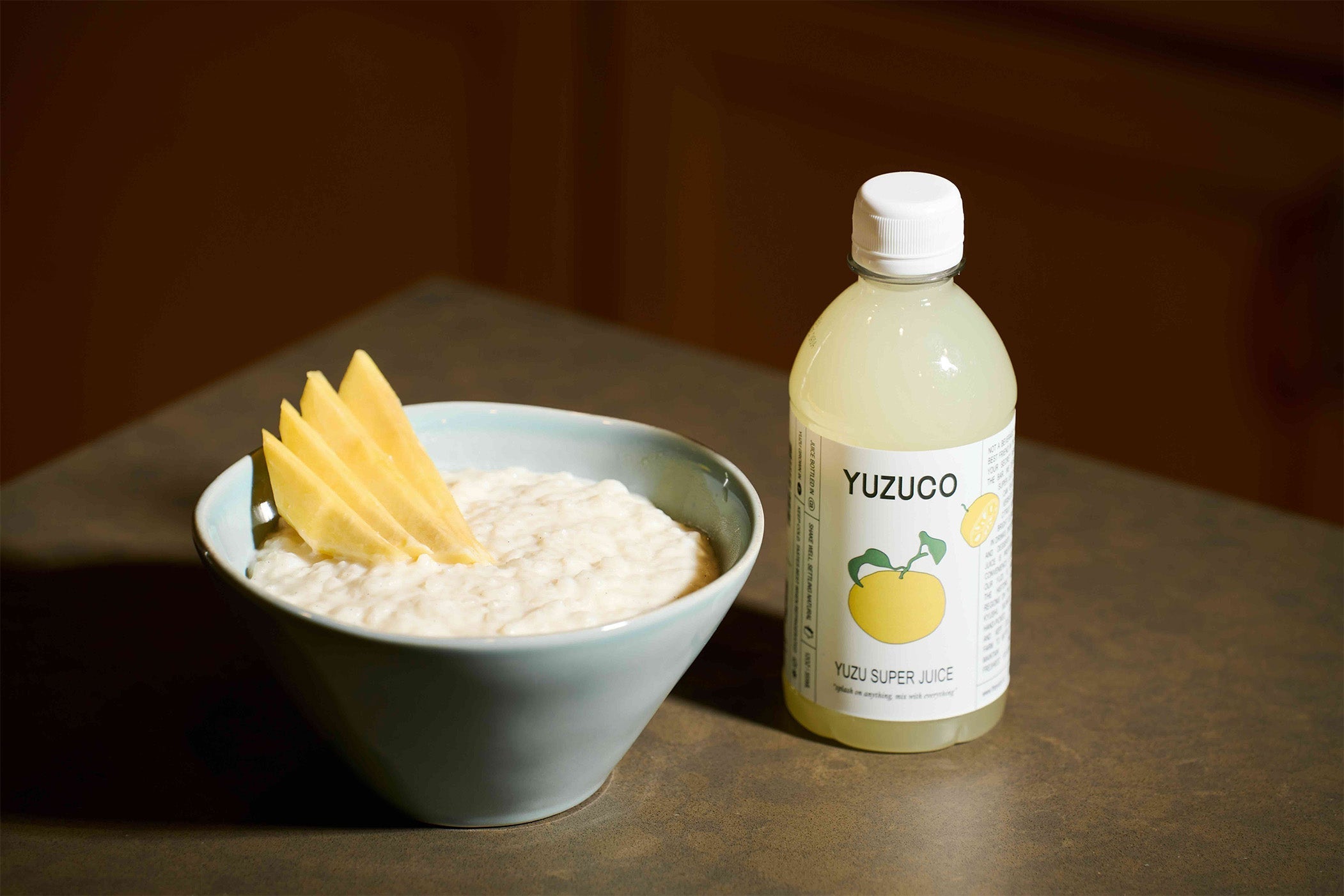 Bowl of yuzu rice pudding topped with sliced mango beside a YUZUCO Yuzu Super Juice bottle on a countertop