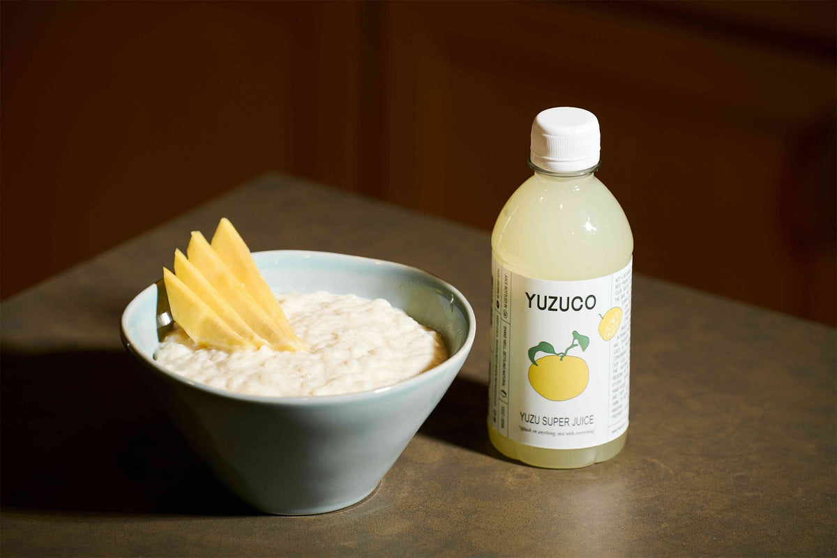 Bowl of yuzu rice pudding topped with sliced mango beside a YUZUCO Yuzu Super Juice bottle on a countertop