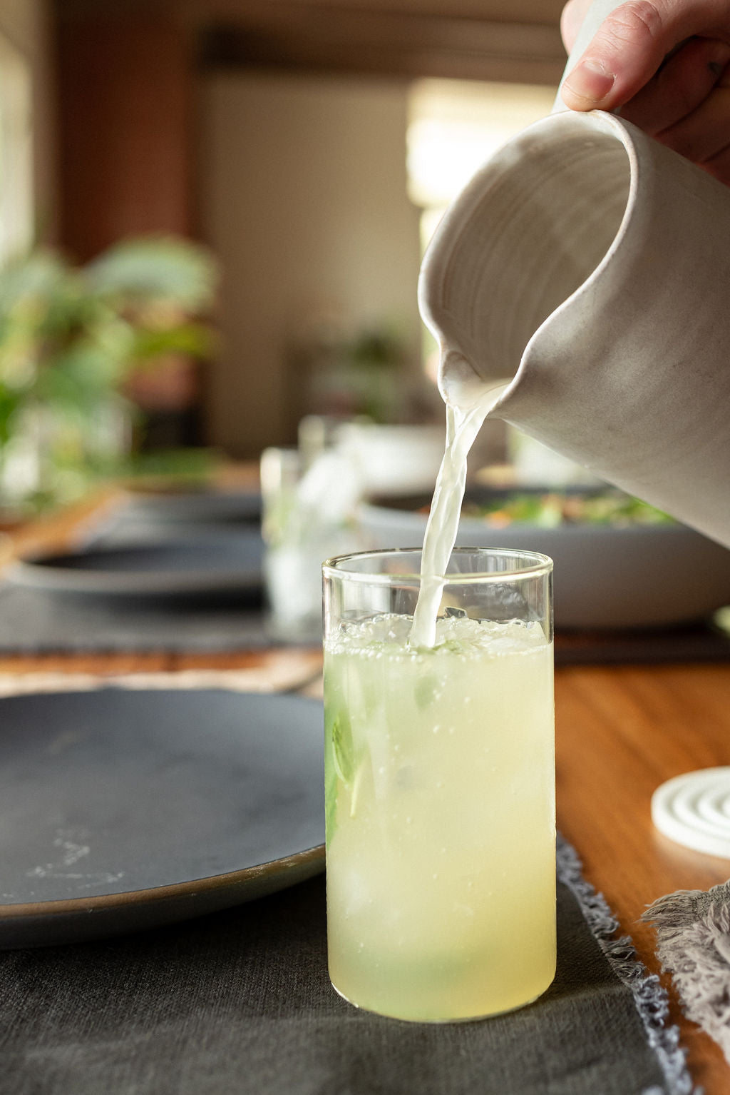 Hand pouring a pale yuzu beverage from a ceramic pitcher into a tall glass with ice on a dining table