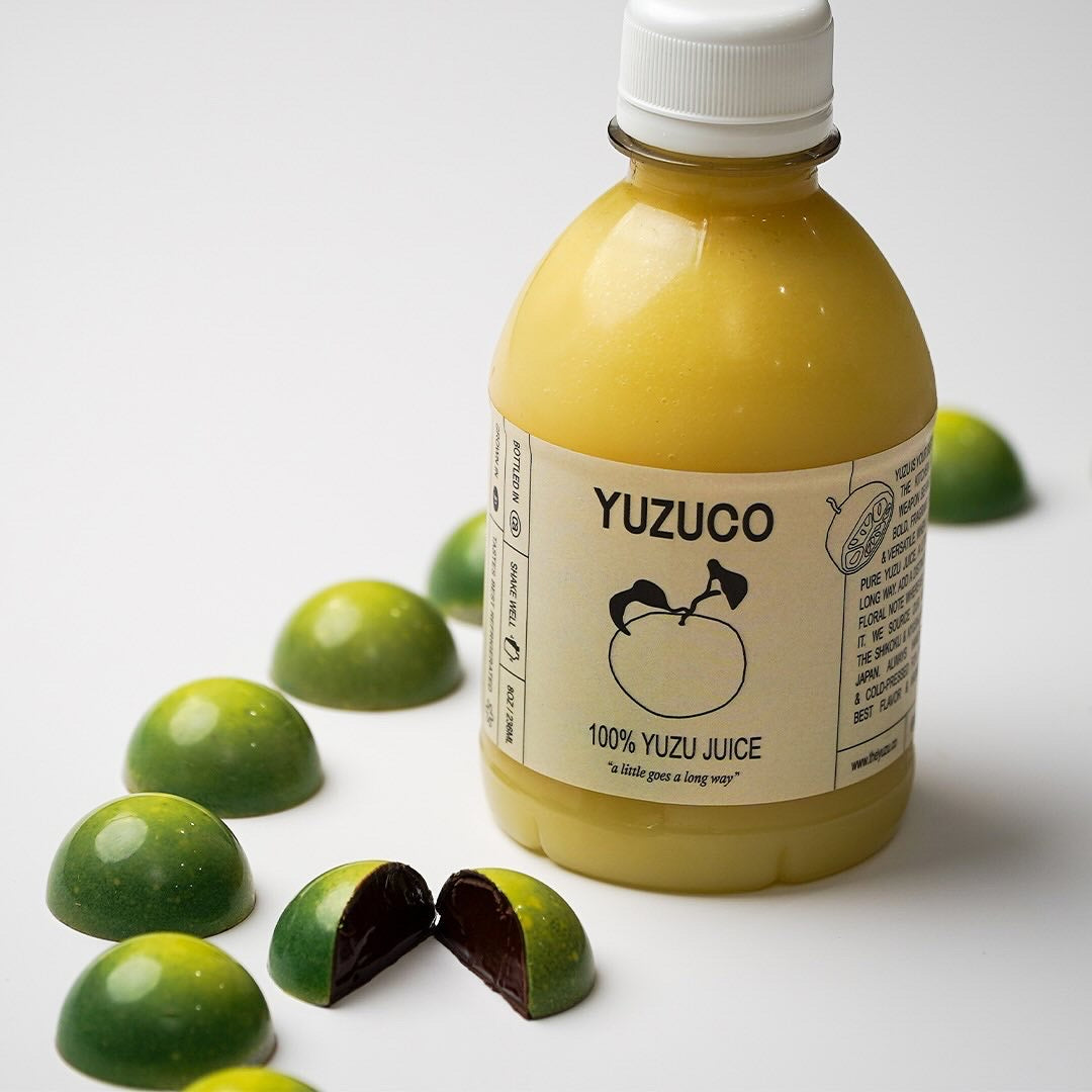 Close-up of yuzu dark chocolate bonbons with glossy green shells and dark ganache beside a bottle of YUZUCO yuzu juice