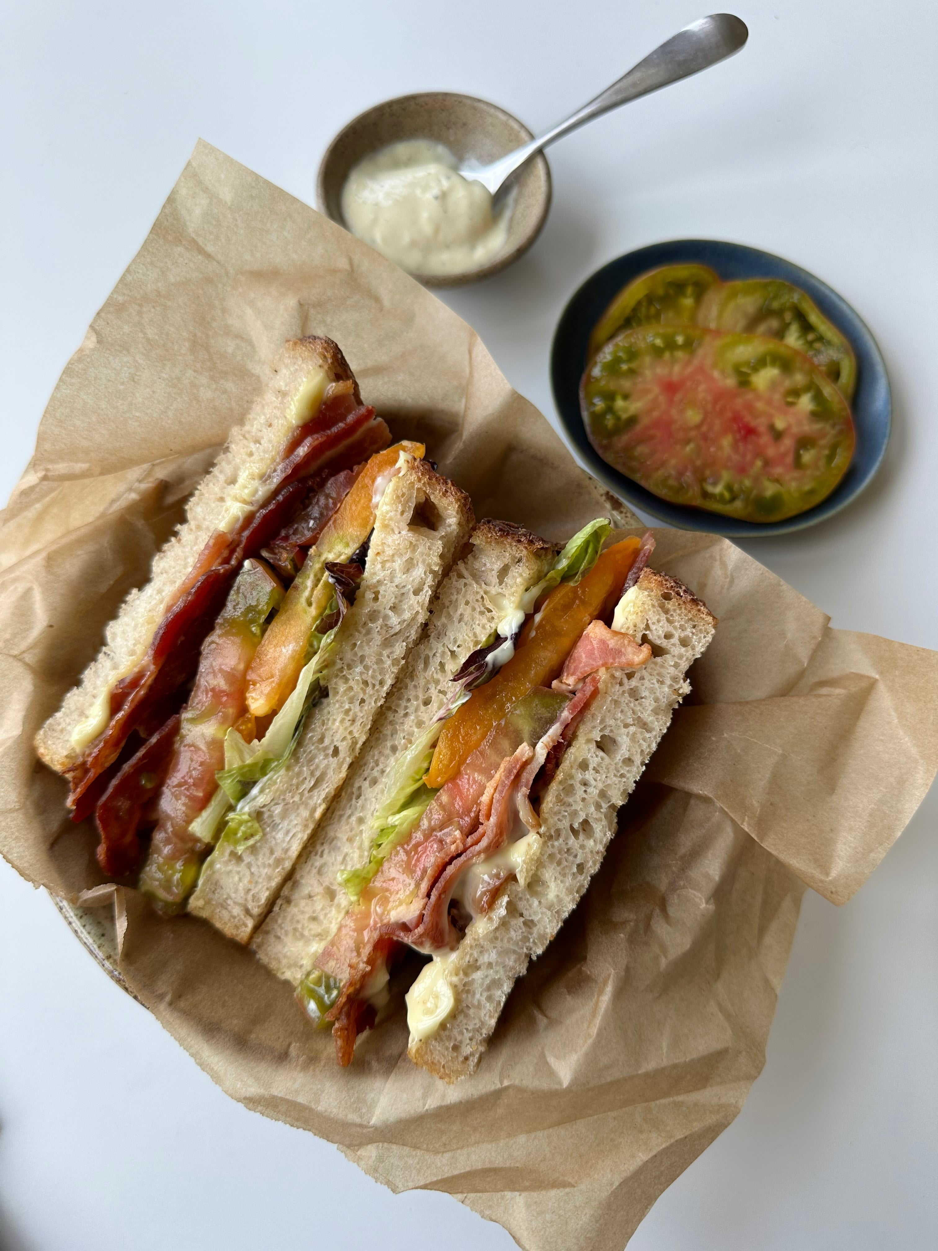 Three BLT halves with bacon, heirloom tomato, and lettuce on sourdough, shown on parchment with a bowl of yuzu aioli