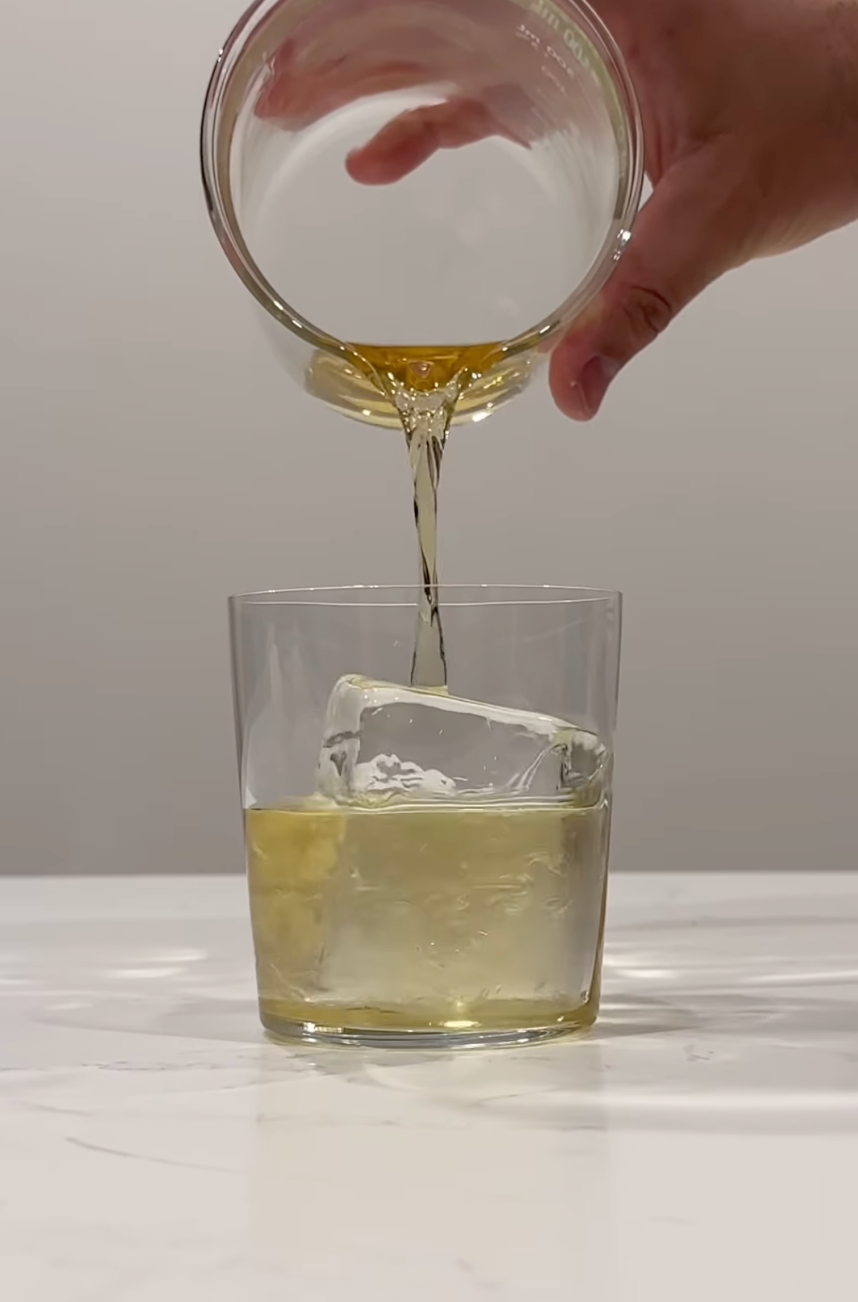 Clarified Yuzu Mai Tai being poured into a rocks glass over a single large ice cube on a white countertop