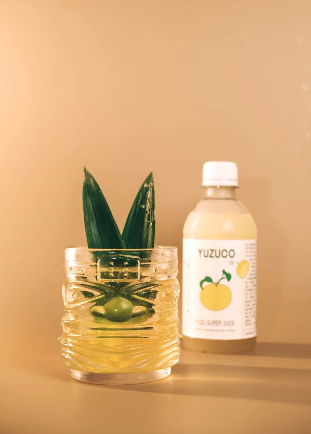 Clarified yuzu Jungle Bird cocktail in a textured glass with pineapple frond garnish beside a YUZUCO bottle on warm beige