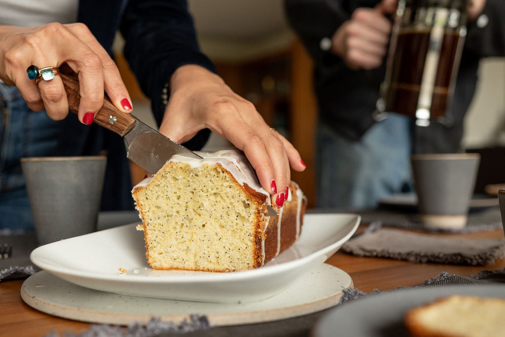 Hands slicing a glazed yuzu poppy seed pound cake on a white platter with coffee cups in the background