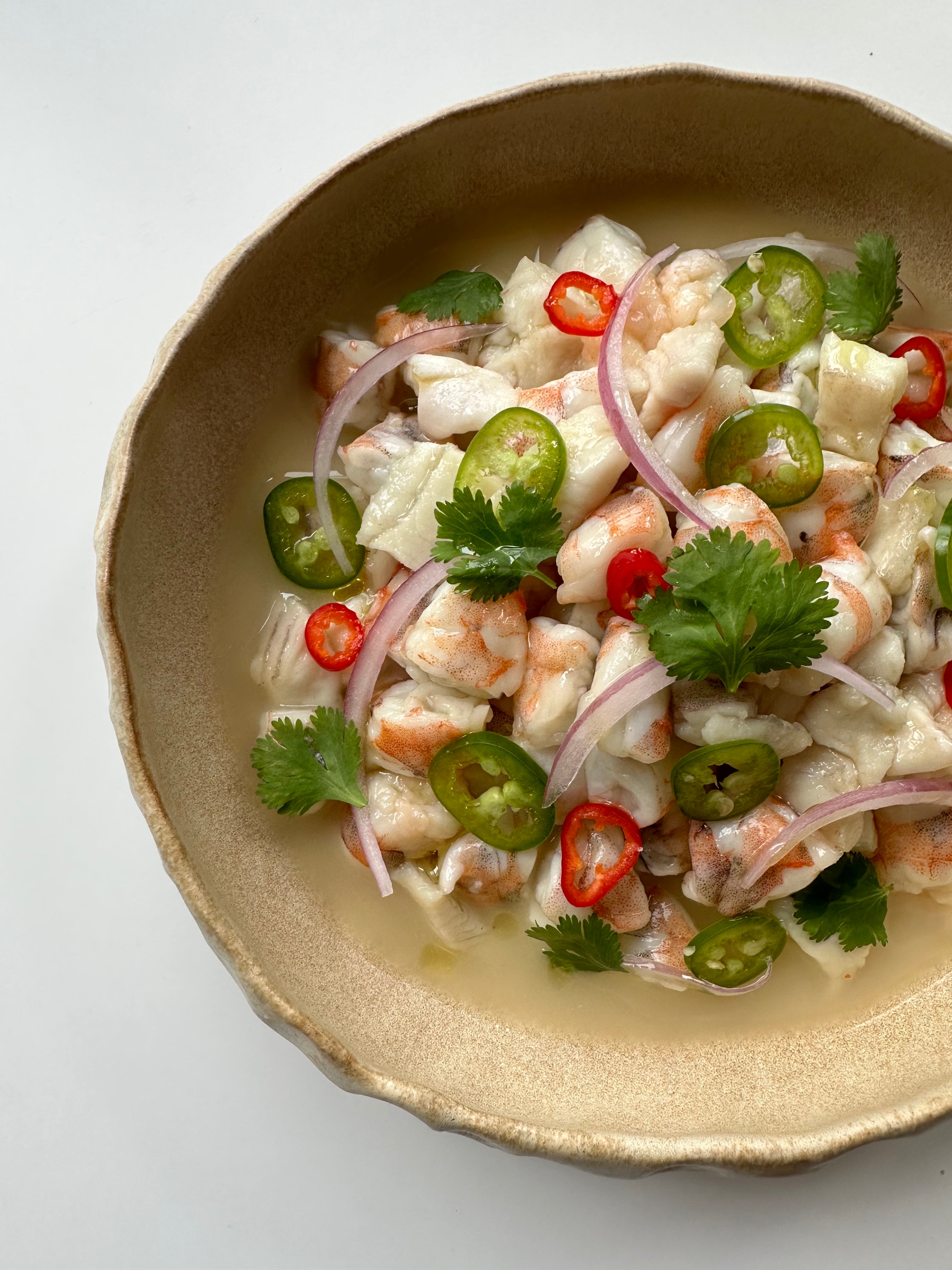 Top-down view of Yuzu ceviche with shrimp and flounder chunks, sliced red onion, red and green chiles, and cilantro in a beige bowl
