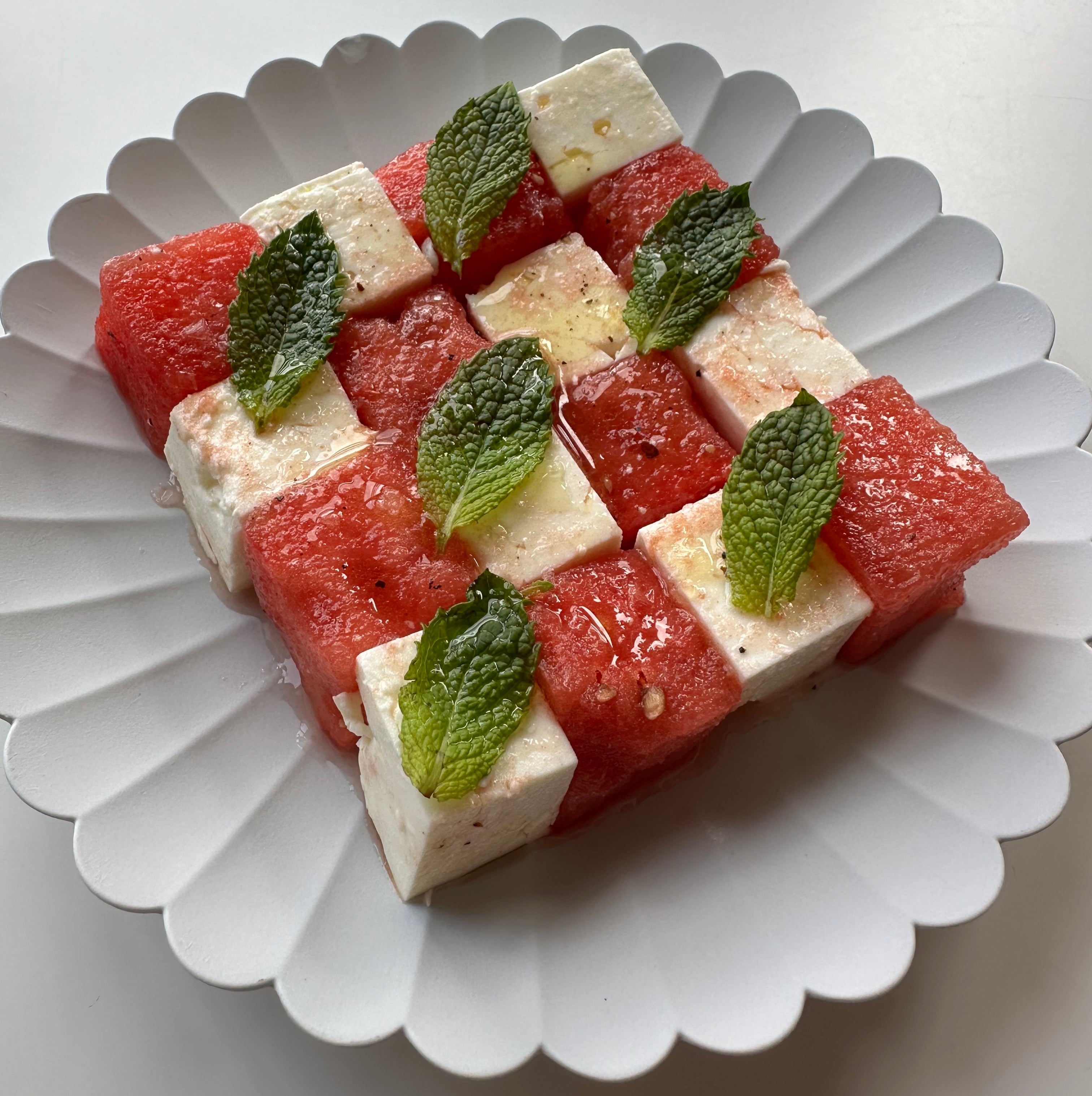 Top-down view of watermelon and feta salad with checkered cubes, mint leaves, and olive oil drizzle on a scalloped white plate