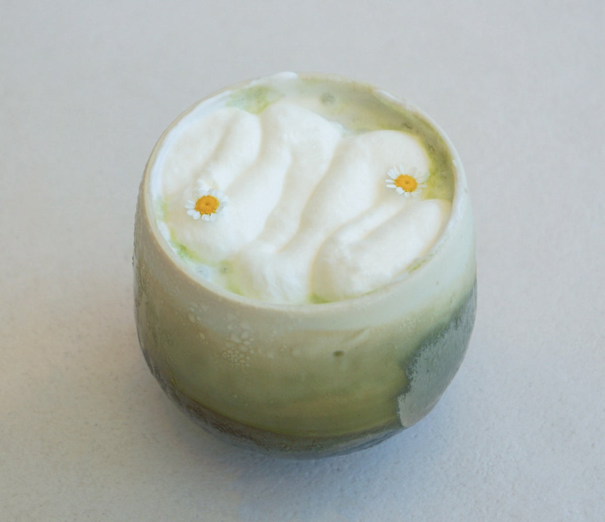 Top view of matcha yuzu cream drink with whipped cream waves and two chamomile flowers in a green cup on pale background
