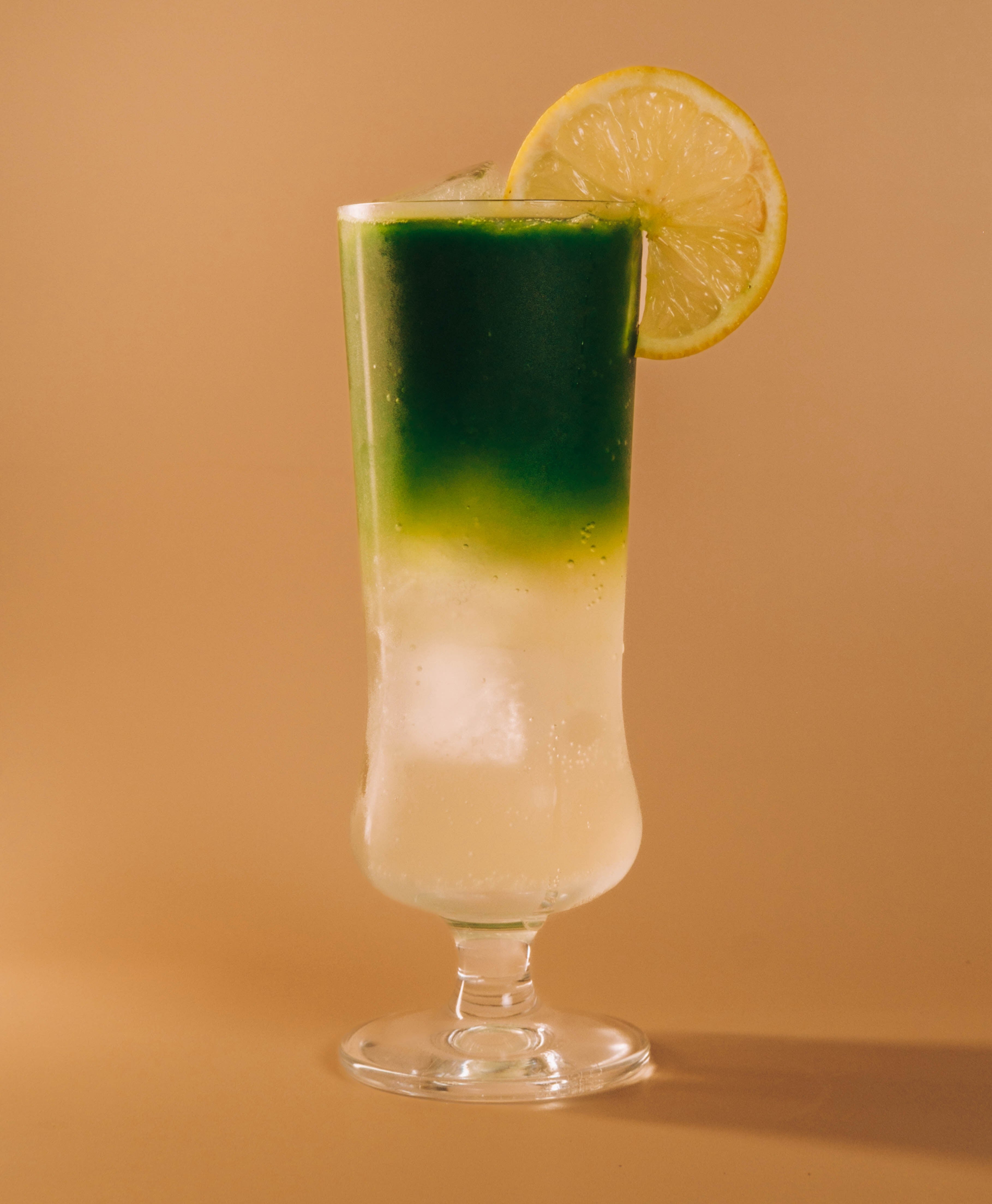 Front view of Yuzu matcha cocktail with layered green matcha over pale lemonade, lemon wheel garnish on a beige background