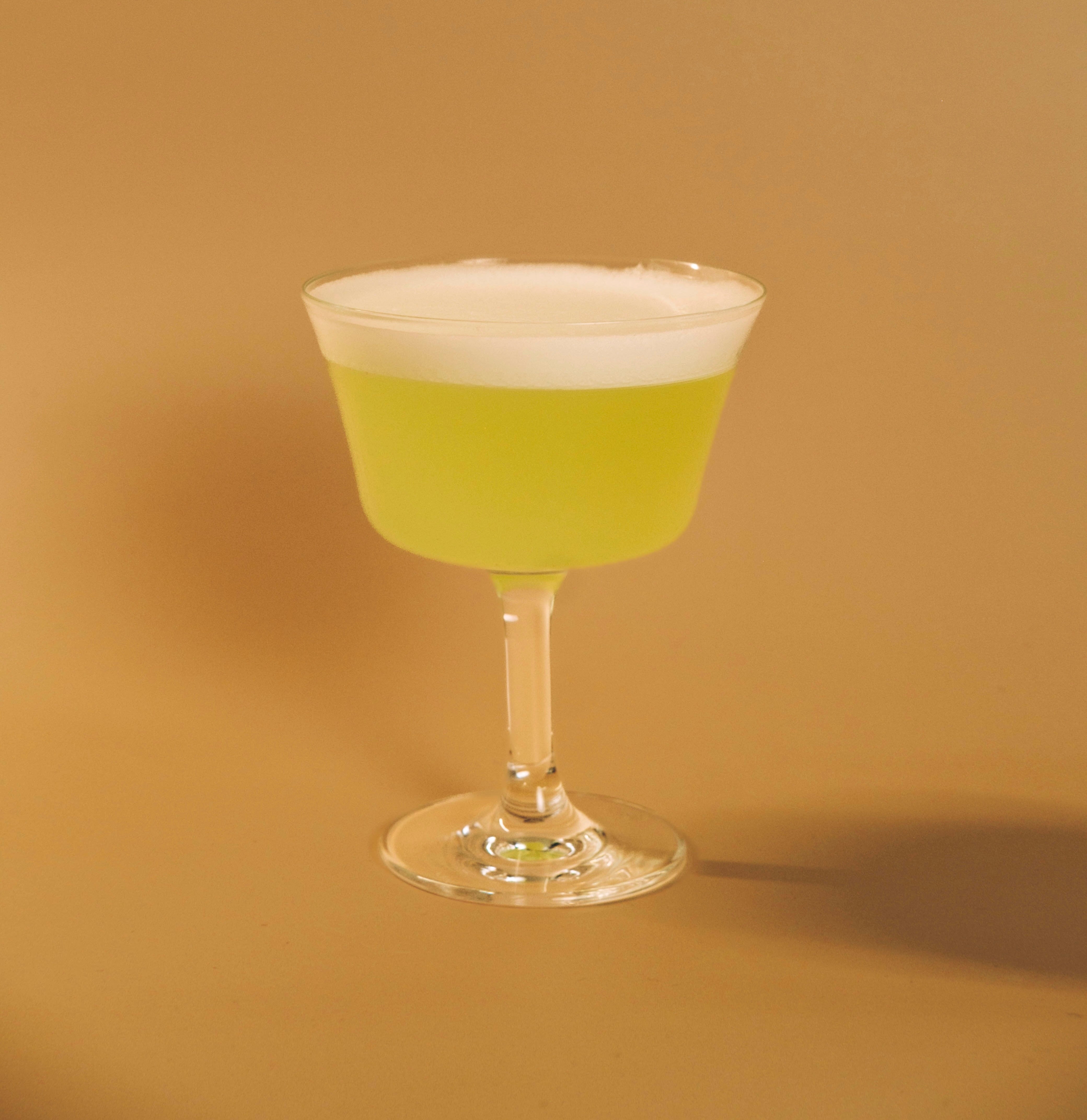 Front view of Sudachi Melon Sour in a coupe glass showcasing bright green liquid and frothy white foam on a beige background