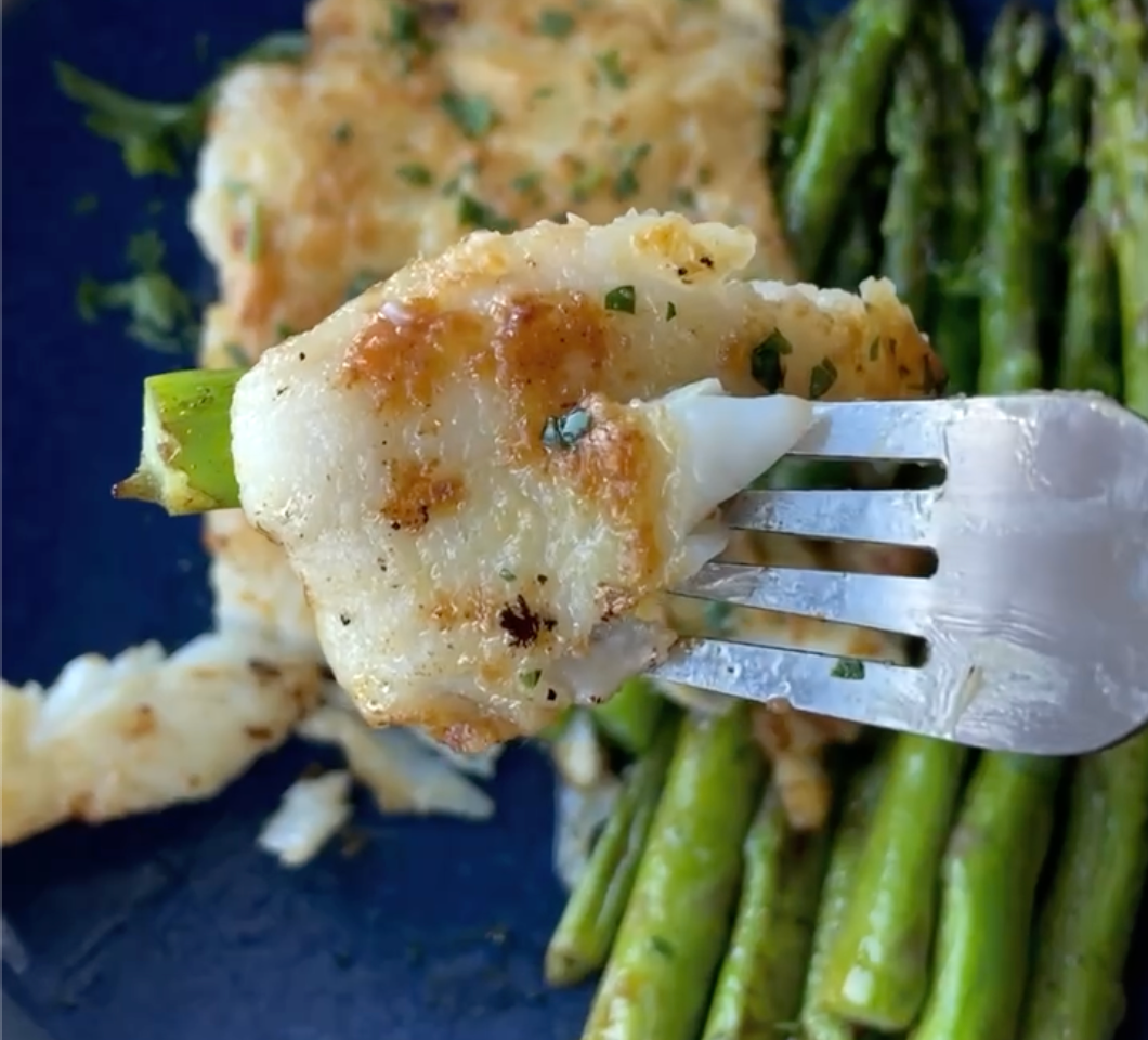 Fork holding a bite of pan-seared yuzu miso-butter cod with golden-browned crust over sautéed asparagus on a dark plate