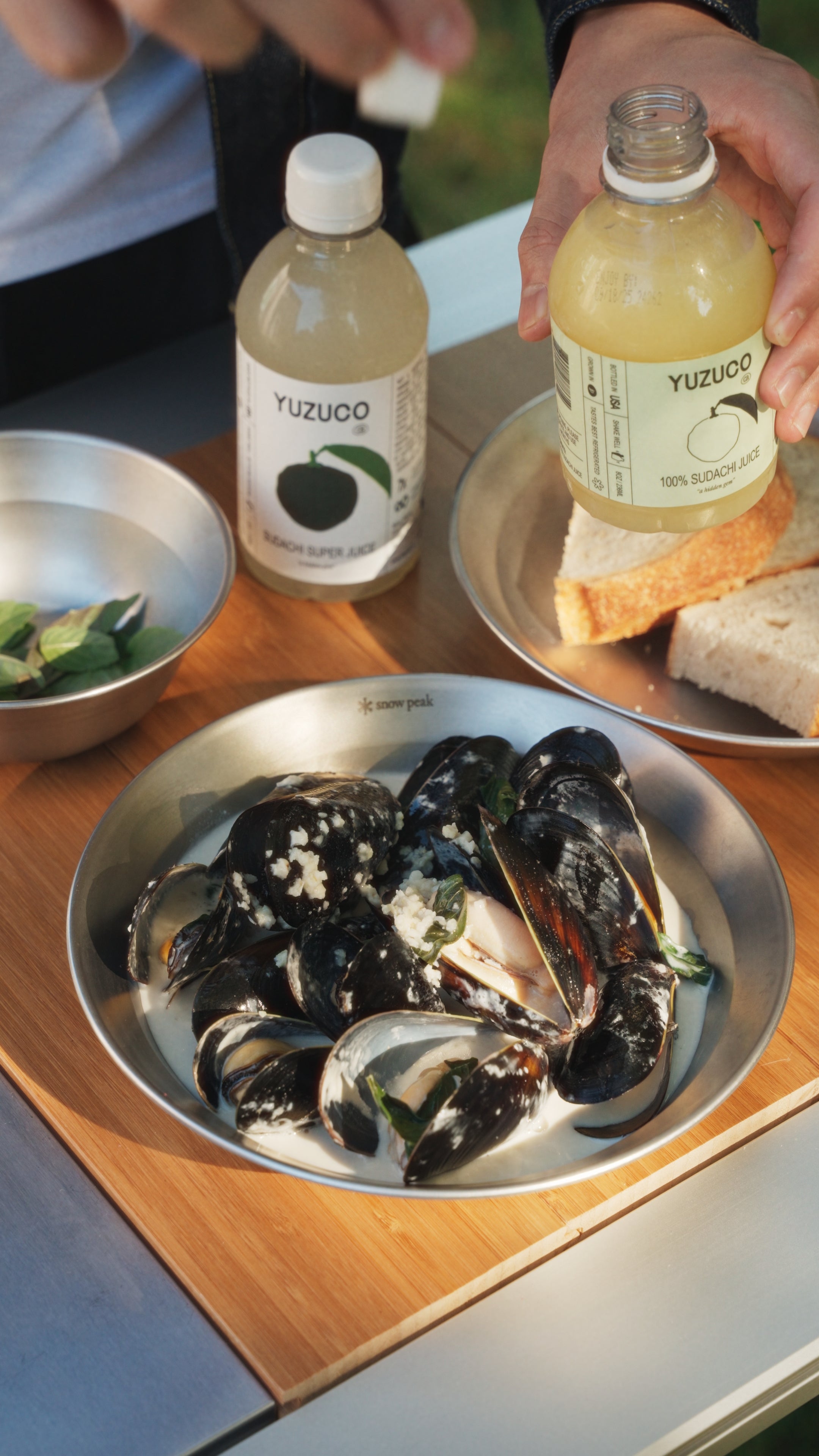 Bowl of steamed mussels in coconut broth with Thai basil, a YUZUCO 100% Sudachi Juice bottle and crusty bread on a wooden table