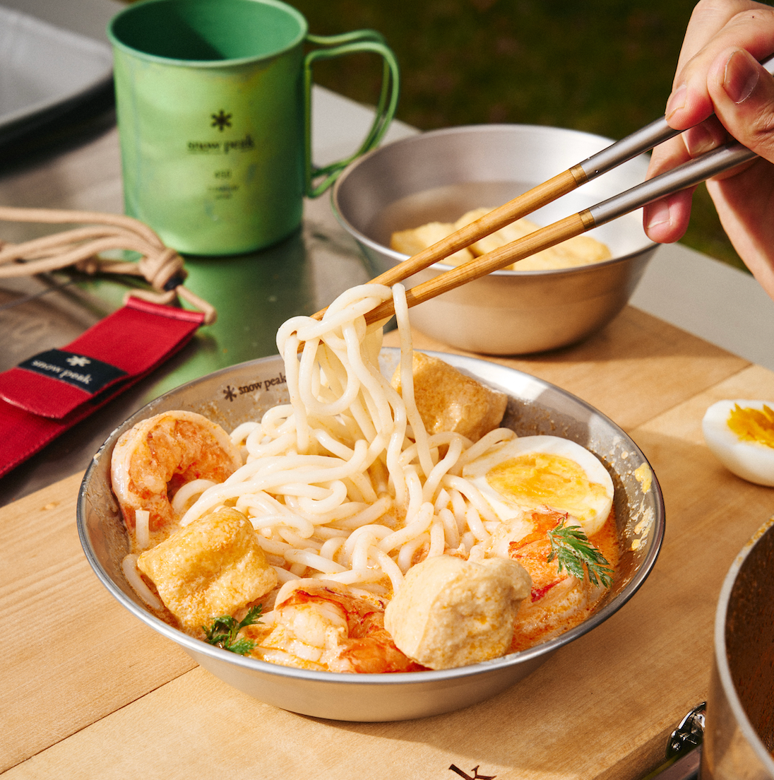 Hand lifting udon noodles from Sudachi curry laksa in a metal bowl with prawns, fried tofu, and halved soft-boiled egg