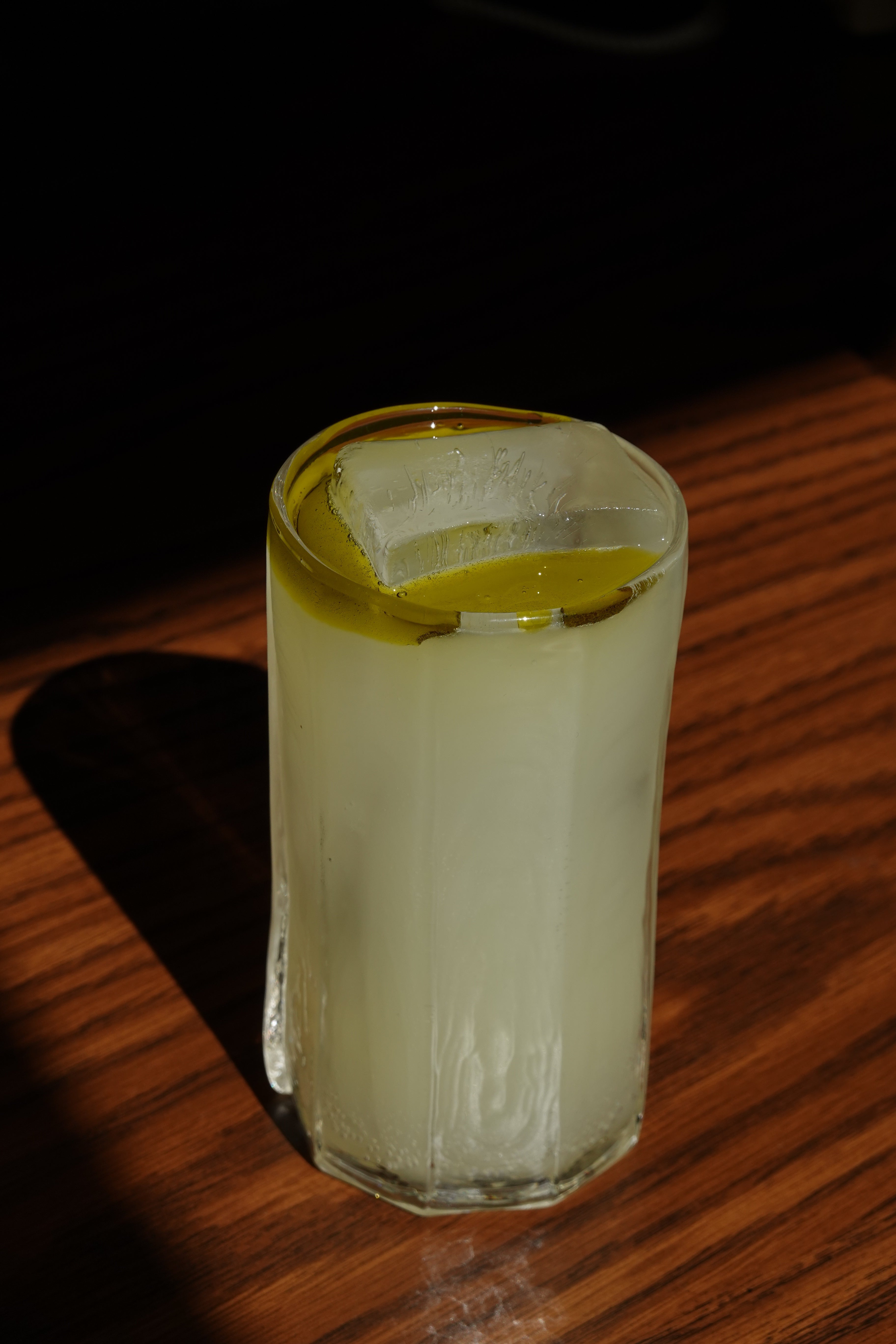 Close-up of Westbourne Mezcalita cocktail in a tall glass showing a large square ice cube and an olive-green oil float on a wood tabletop