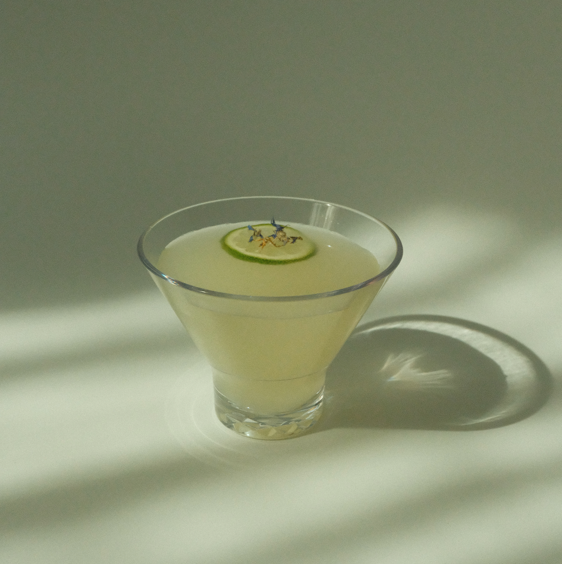 Overhead view of Yuzu Daiquiri in a stemless glass with a lime wheel and tiny edible flowers on a sunlit surface