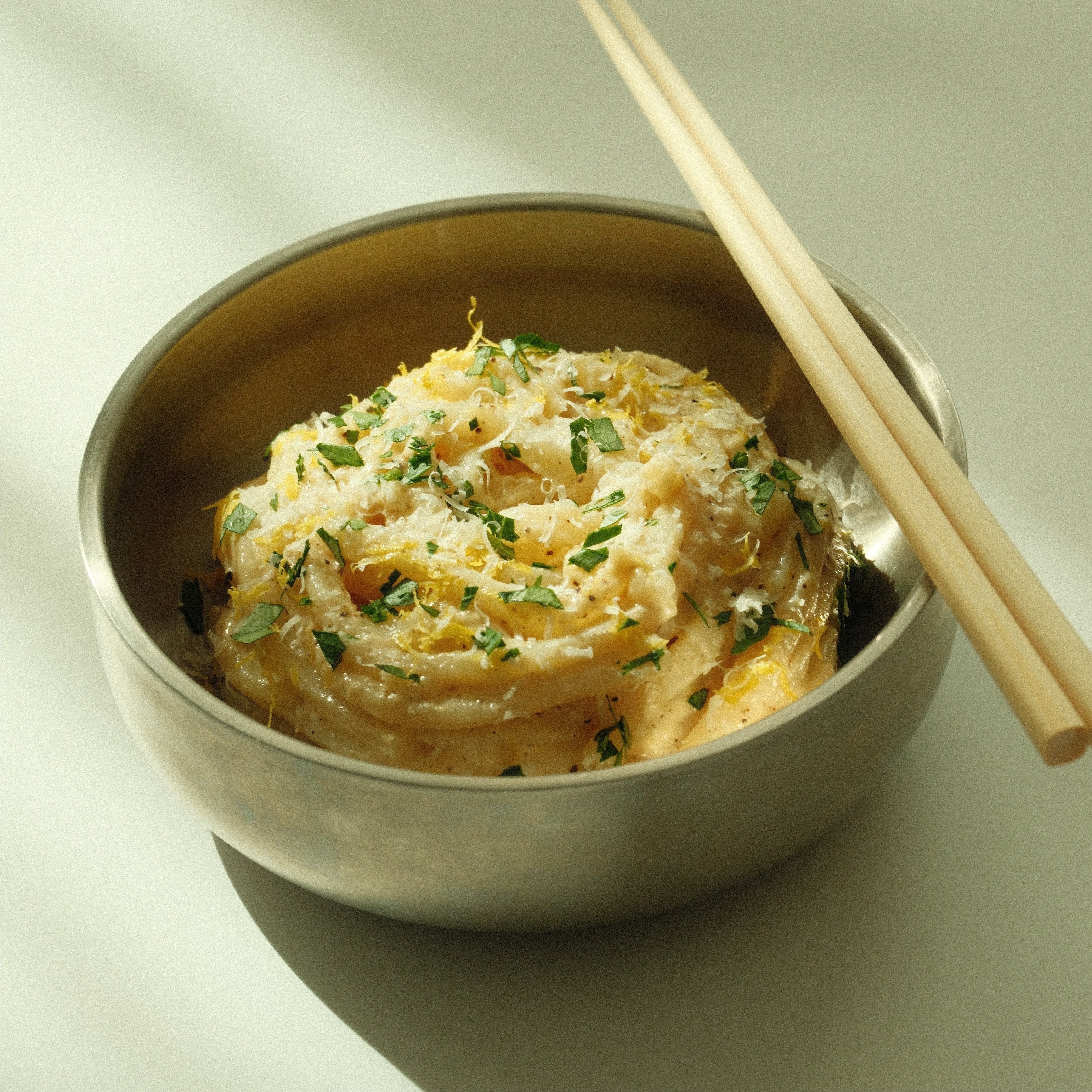 Bowl of creamy yuzu udon noodles topped with grated parmesan, lemon zest, and chopped parsley, chopsticks resting on the rim
