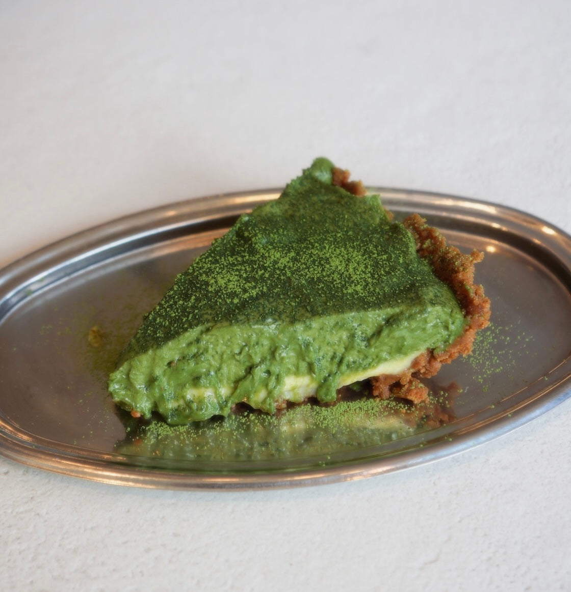 Side view of a slice of matcha mousse pie with creamy green filling and graham cracker crust on a silver oval plate