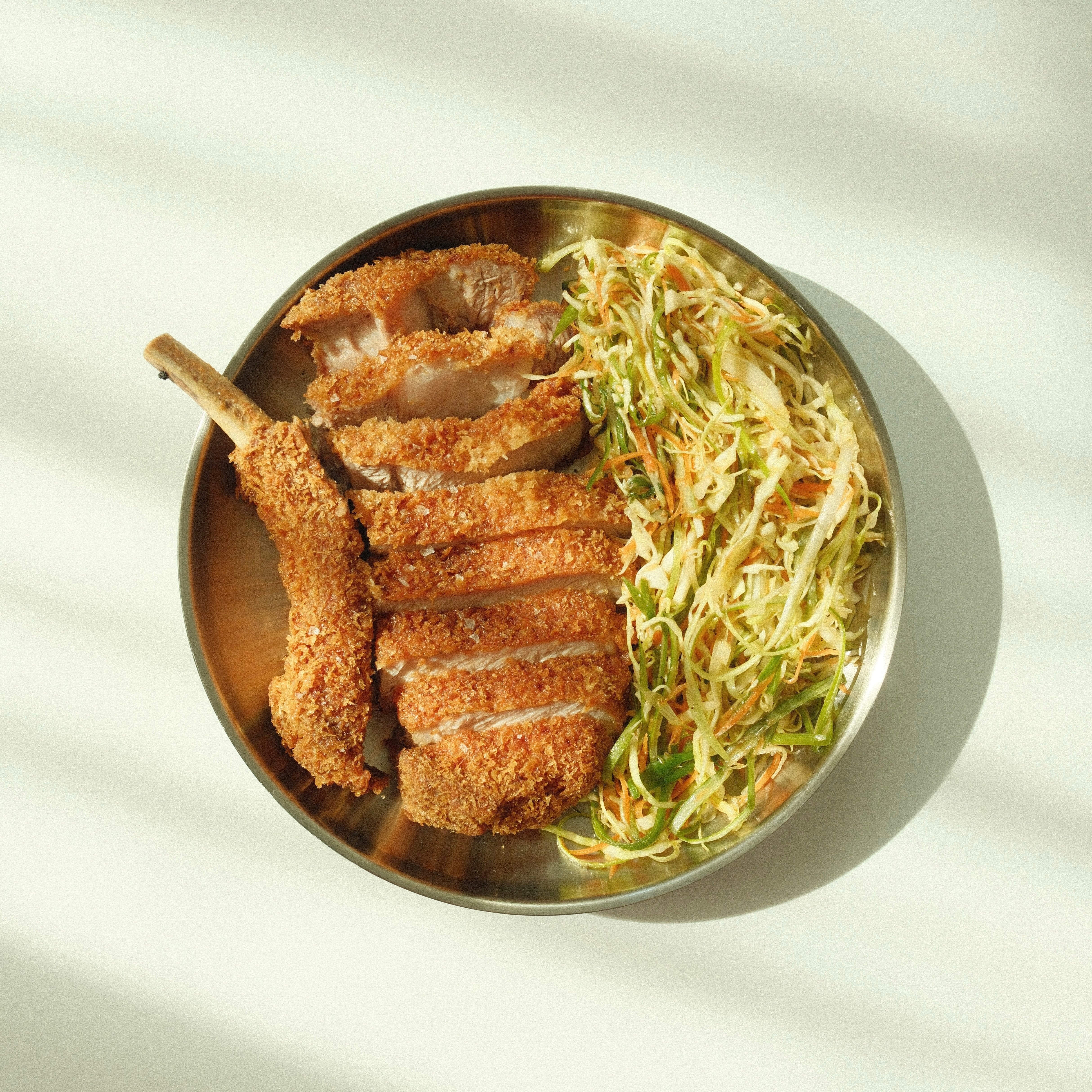 Top-down view of sliced breaded pork cutlet with shredded cabbage slaw in a brass bowl on a pale background