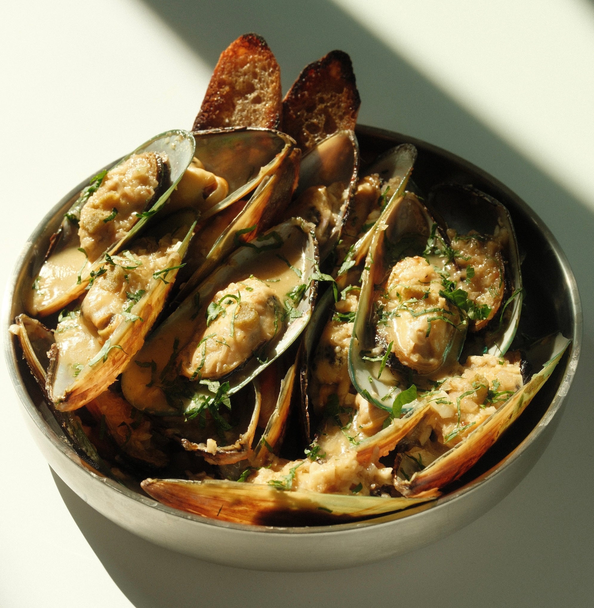 Close-up of yuzu kosho mussels in a buttery sauce, garnished with chopped parsley and served with toasted baguette in a metal bowl