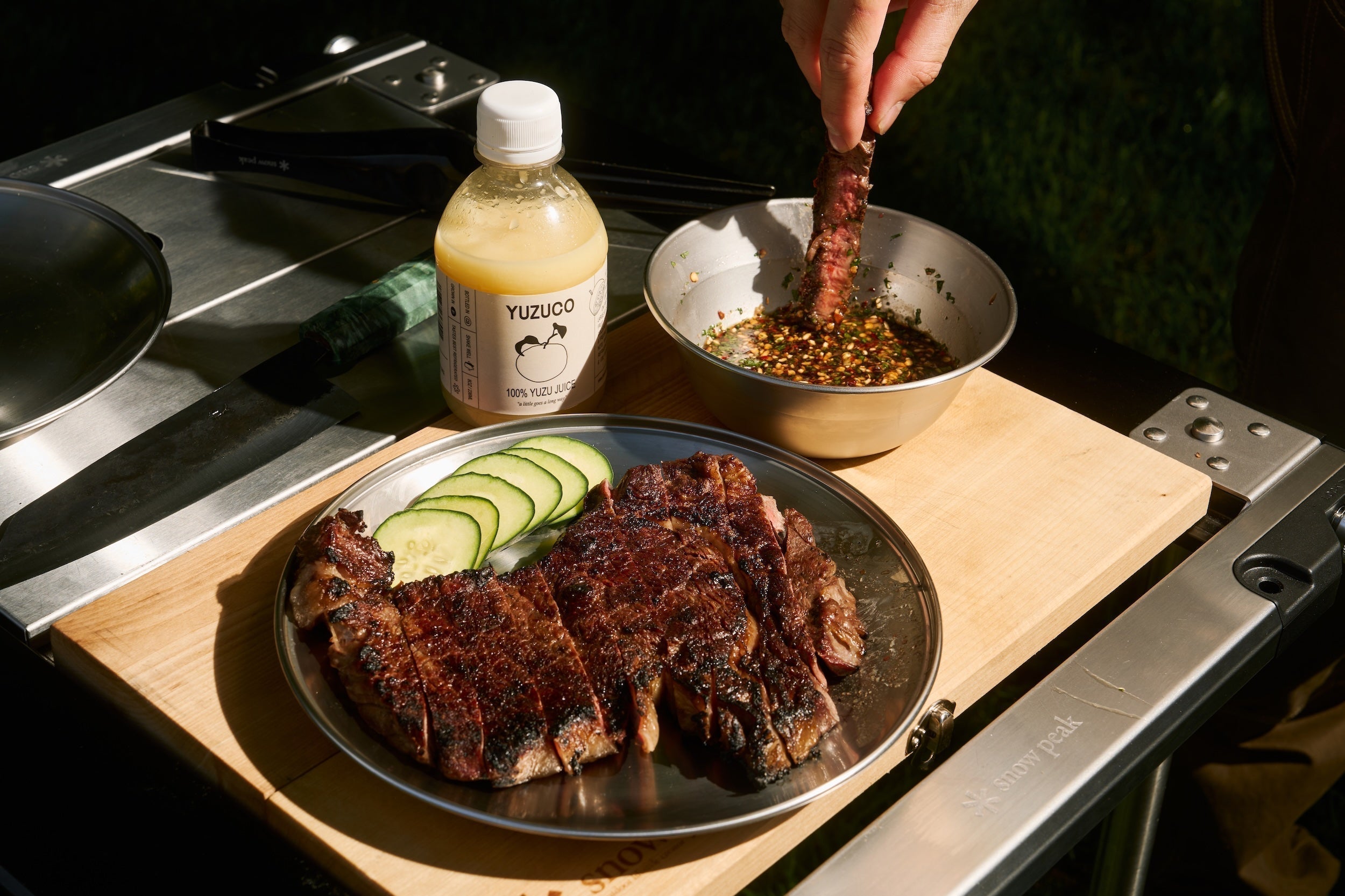 Grilled Crying Tiger steak dipped into spicy Nam Jim Jaew sauce beside a bottle of YUZUCO yuzu juice on a cutting board