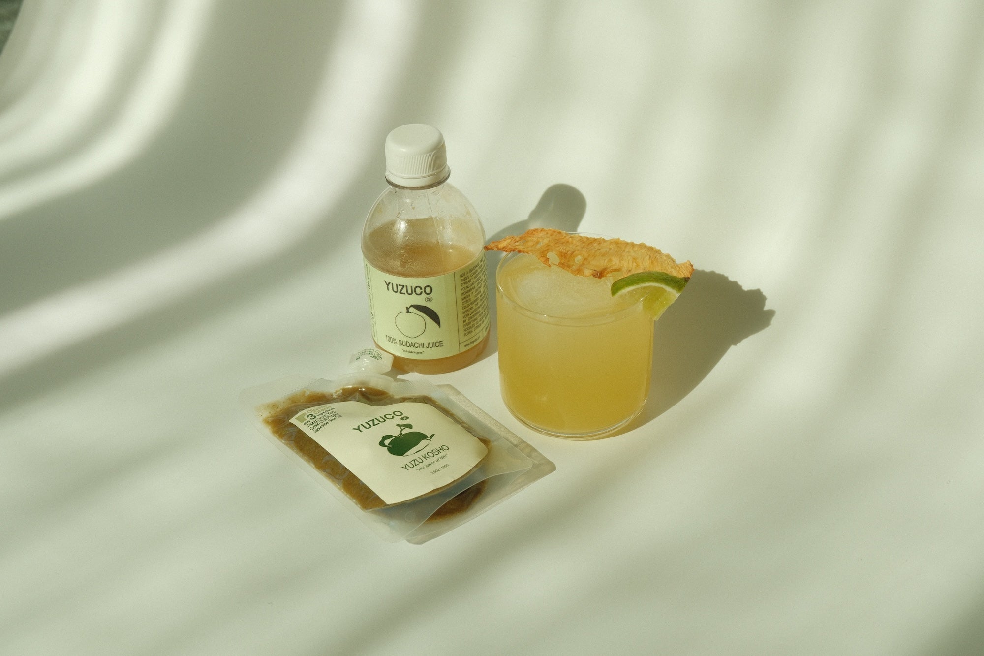 Yuzu kosho margarita cocktail beside a YUZUCO bottle and yuzu kosho packet on a sunlit fabric background