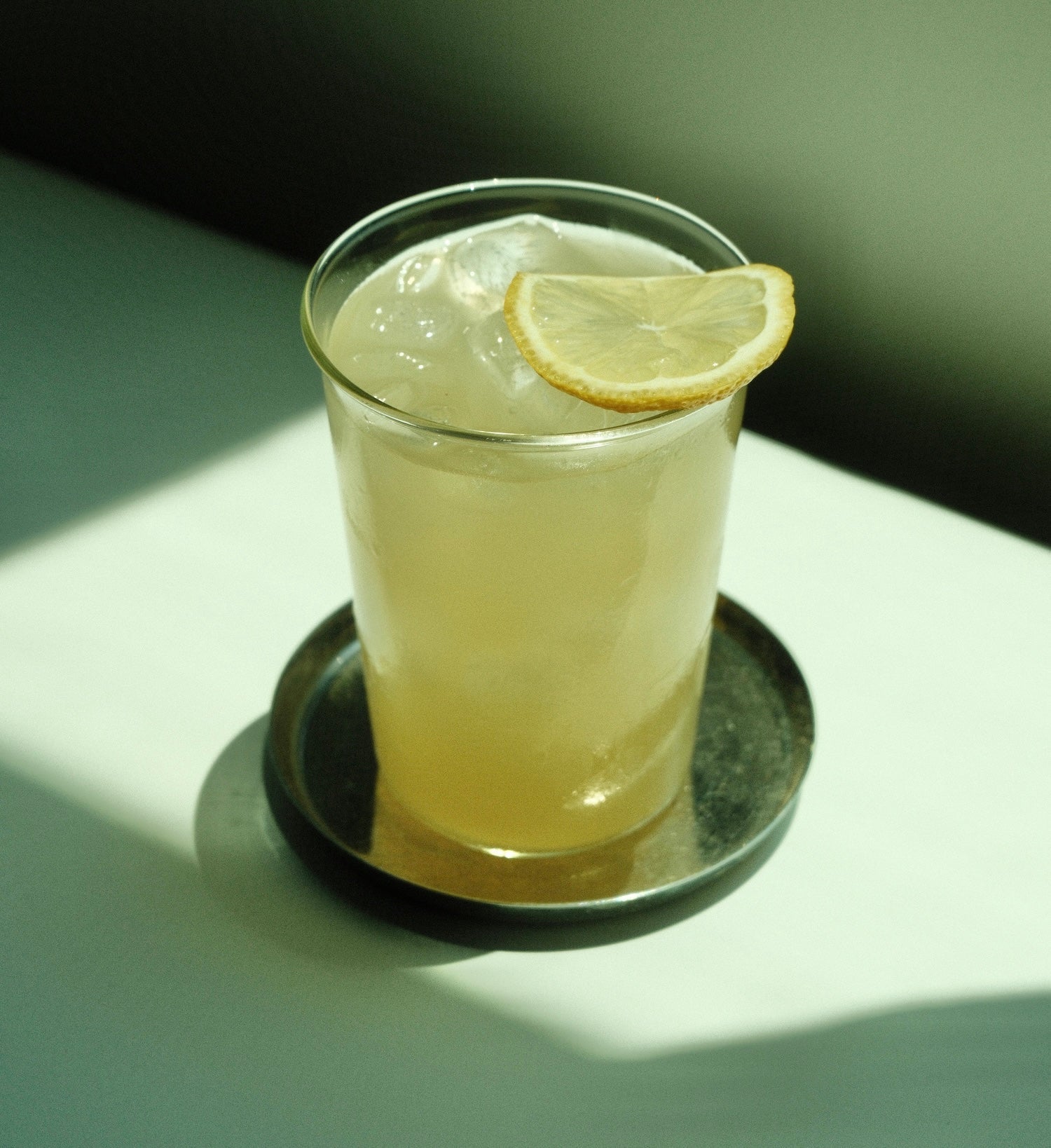 Close up of a tall glass of yuzu ade showing ice and a lemon slice on a metal coaster against a softly lit green background