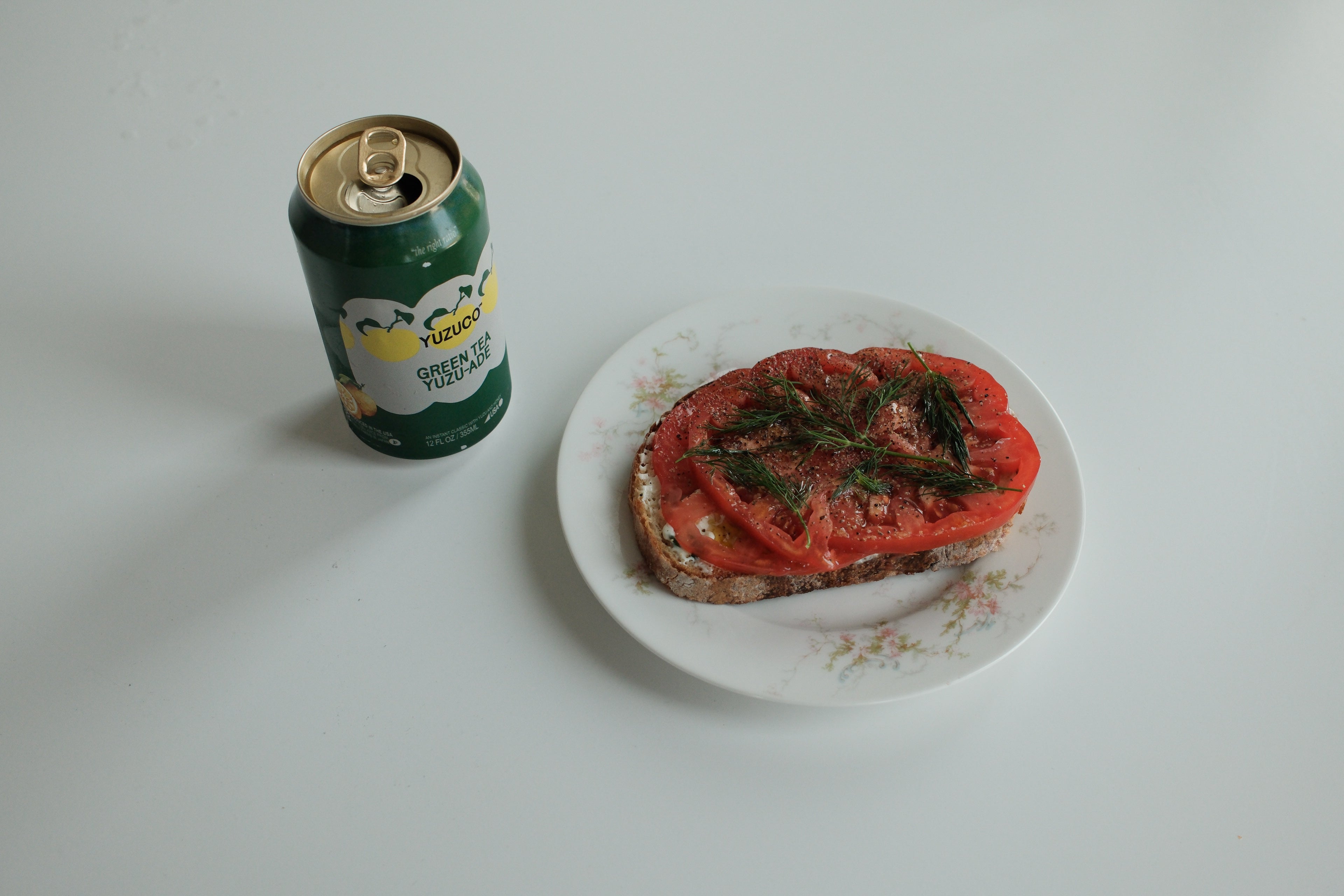 Tomato toast topped with yuzu kosho scallion cream cheese, sliced heirloom tomato, dill and cracked pepper on a floral plate beside a can on a white table