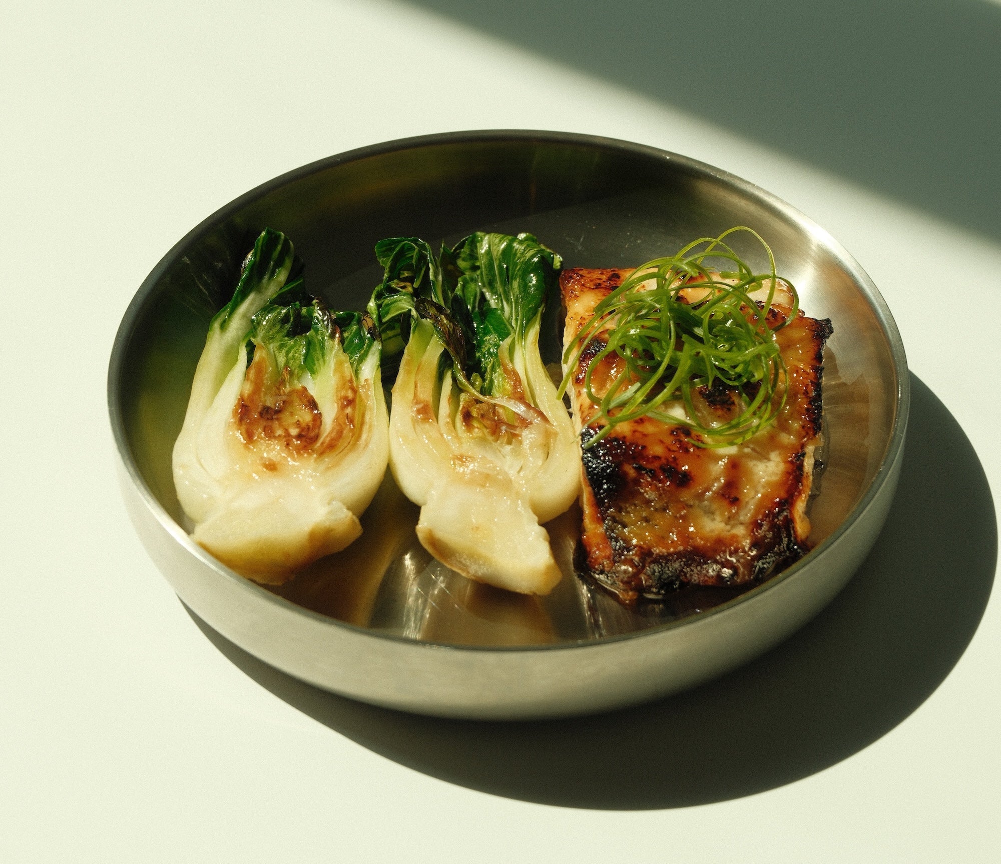 Miso-glazed black cod with charred baby bok choy on a round metal plate, topped with thinly sliced scallions