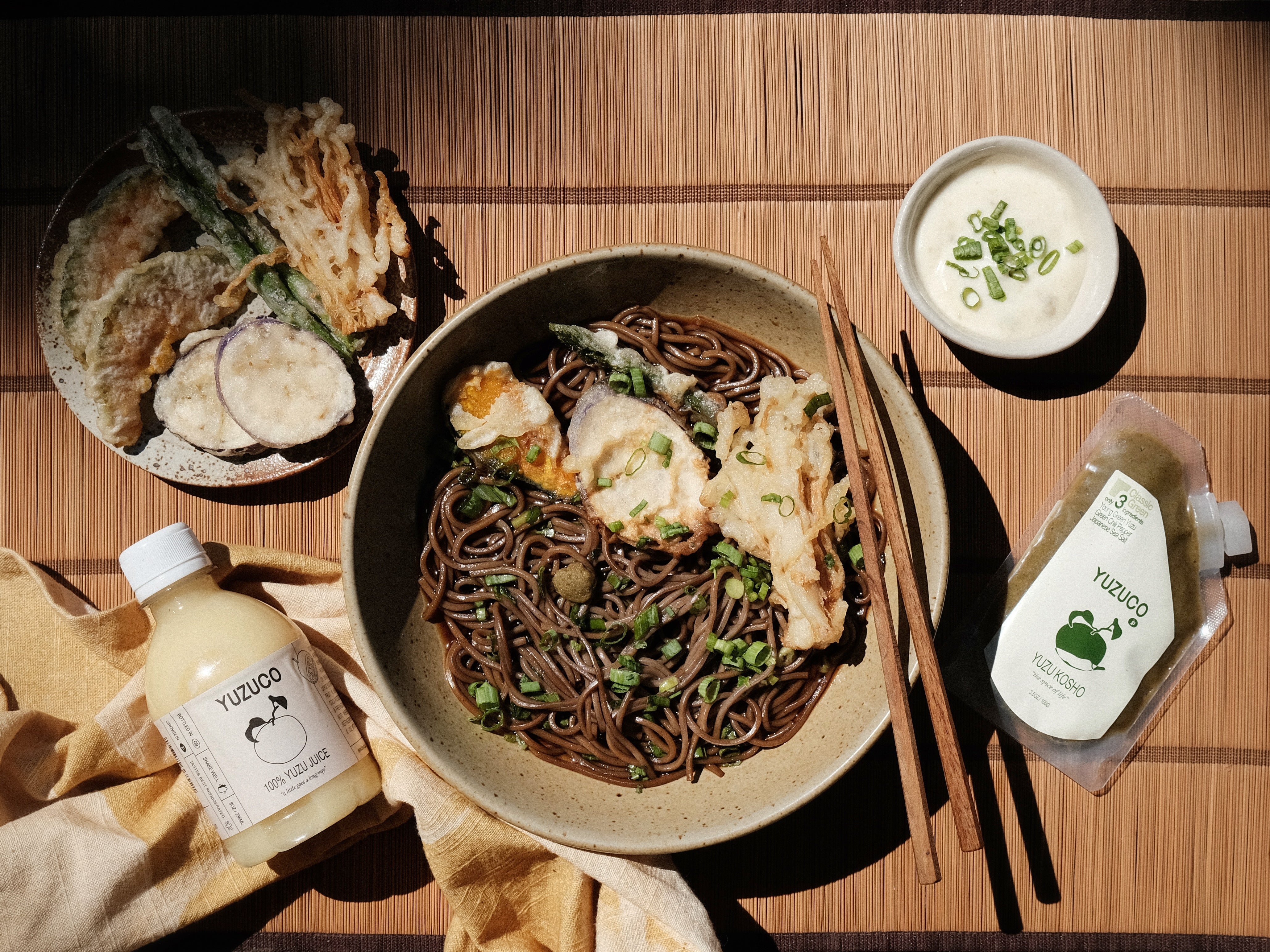 Chilled soba with tempura and scallions on a bamboo mat, with chopsticks and YUZUCO yuzu juice bottle and yuzu kosho pouch
