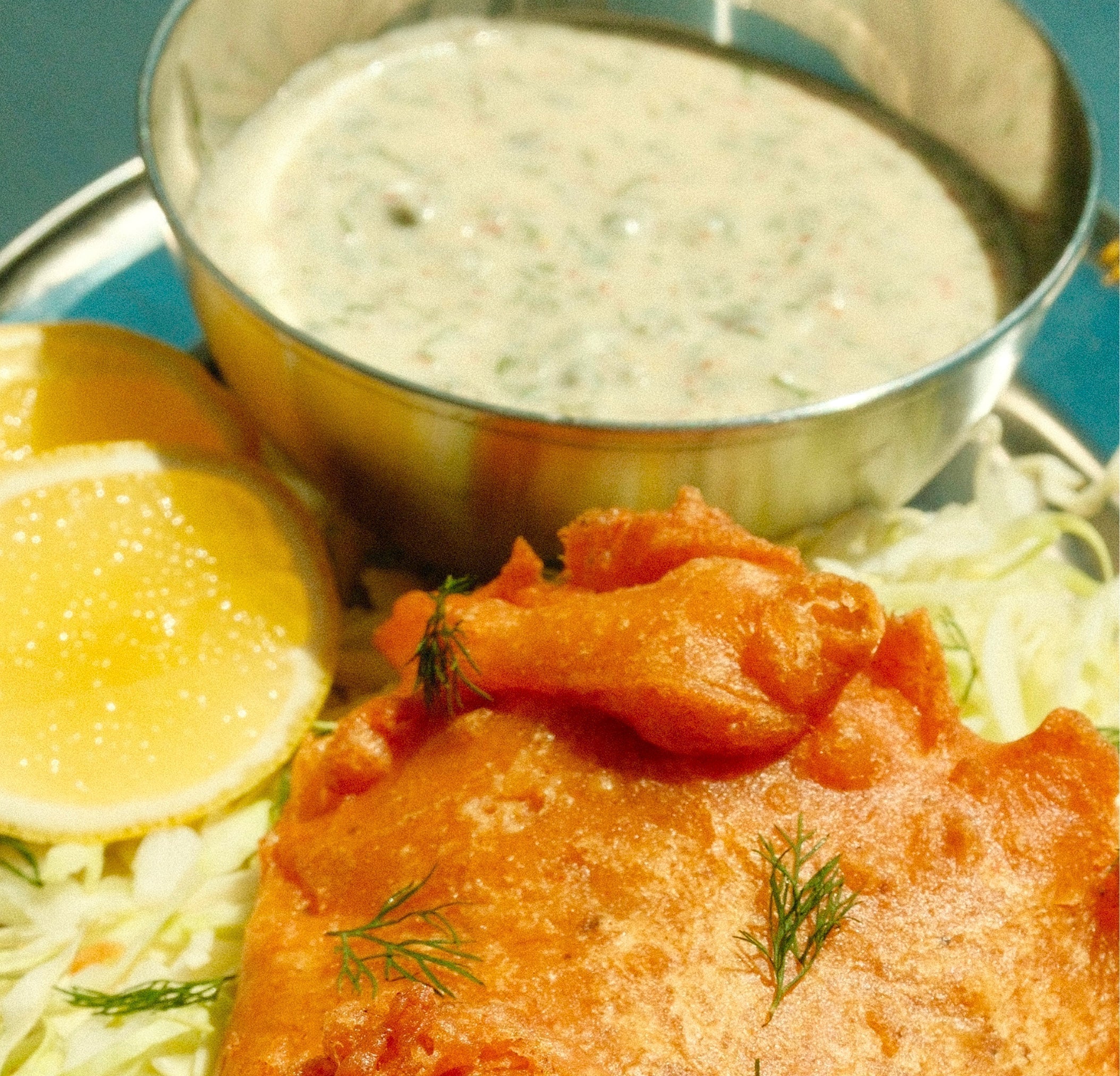 Close up of yuzu tartar sauce in a metal ramekin showing creamy texture and herb flecks with lemon wedges and fried fish