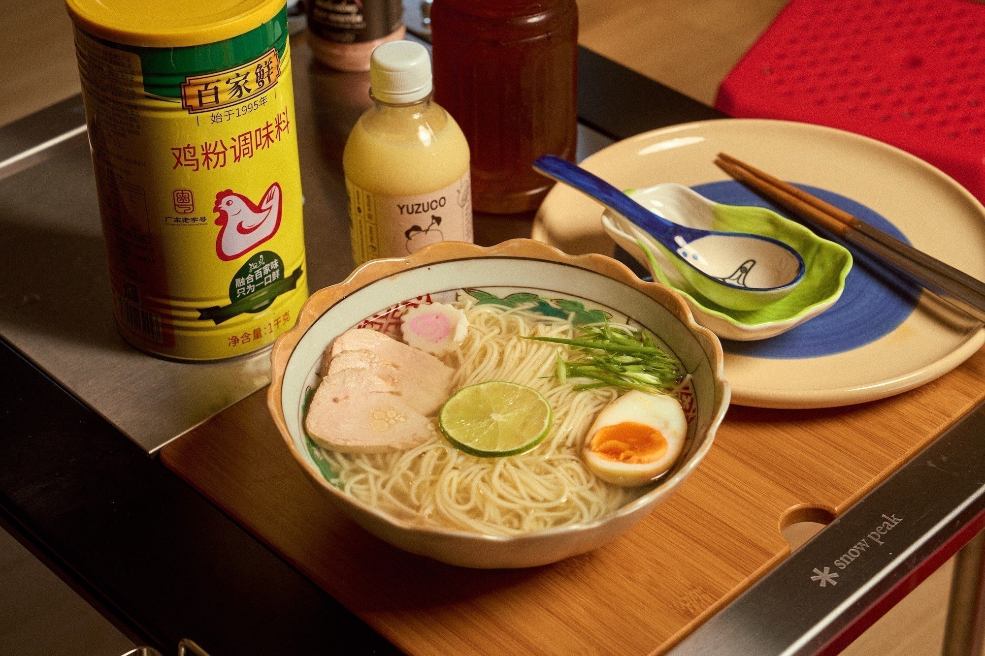 Bowl of yuzu chicken ramen with sliced chicken, soft-boiled egg and lime on a wooden board beside a YUZUCO bottle and chopsticks