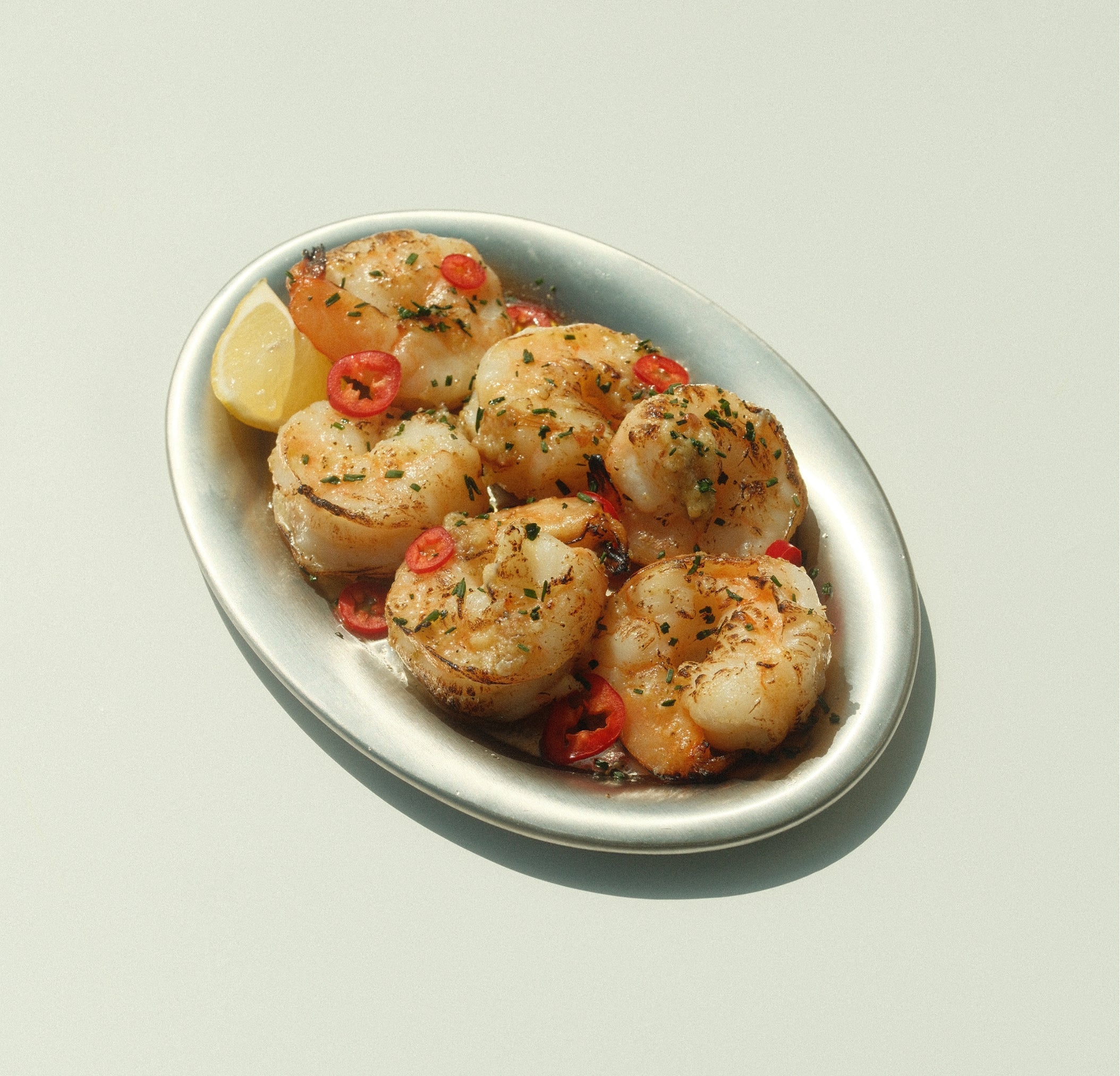 Seared shrimp tossed in yuzu kosho compound butter on a metal oval plate with sliced Fresno chiles, chives, and lemon wedge