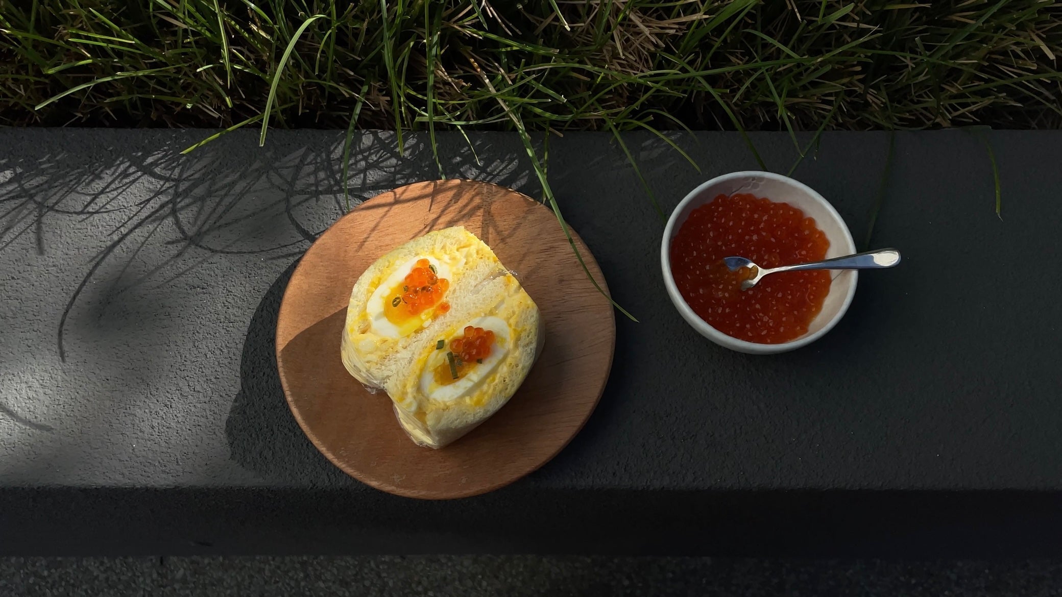 Top-down view of yuzu egg mentaiko sando halved on a wooden plate, topped with ikura and chives beside a bowl of salmon roe on a dark stone ledge