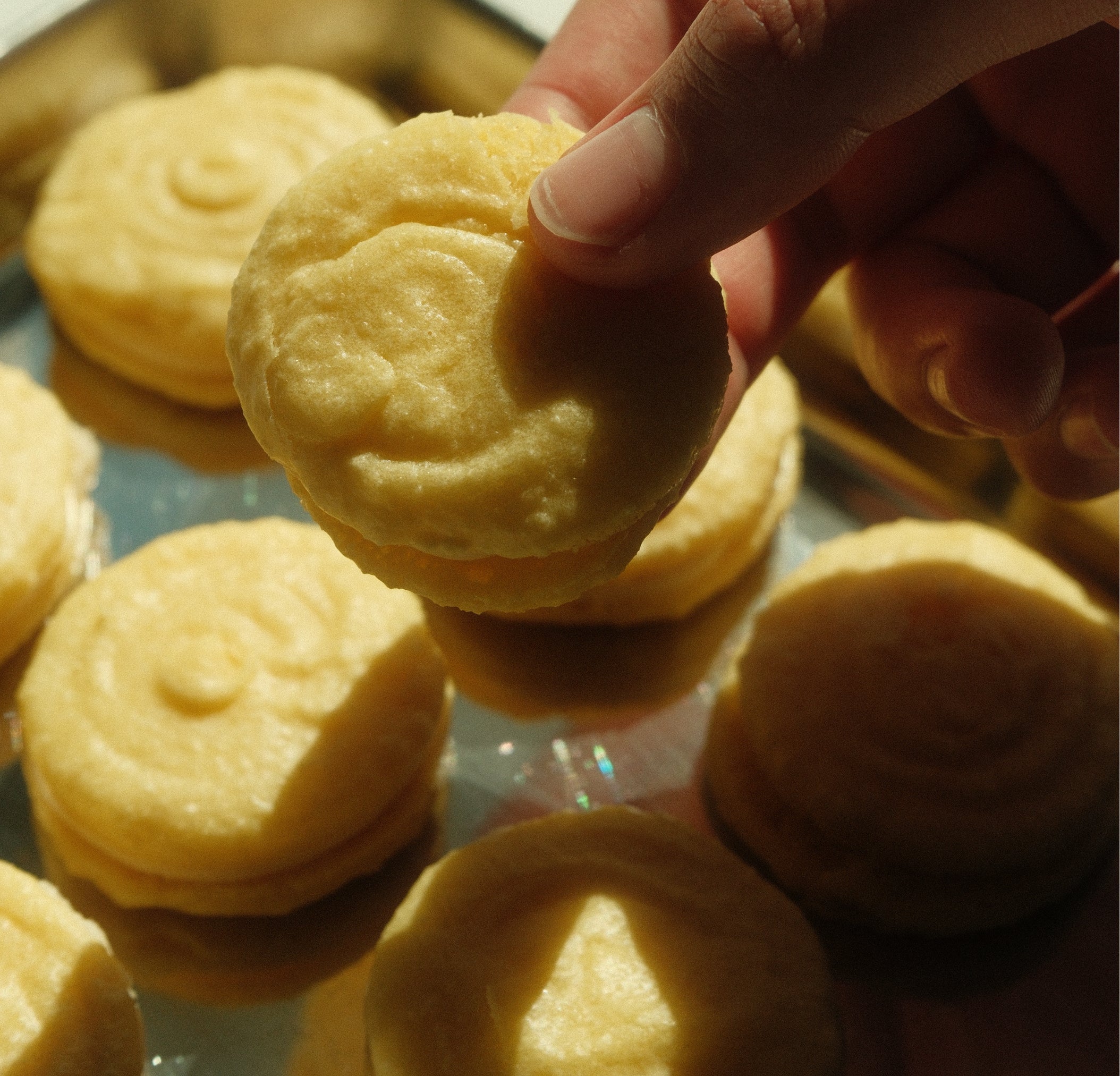 Close-up of pale yellow yuzu macaron held between fingers showcasing smooth shell and buttercream filling on shiny tray