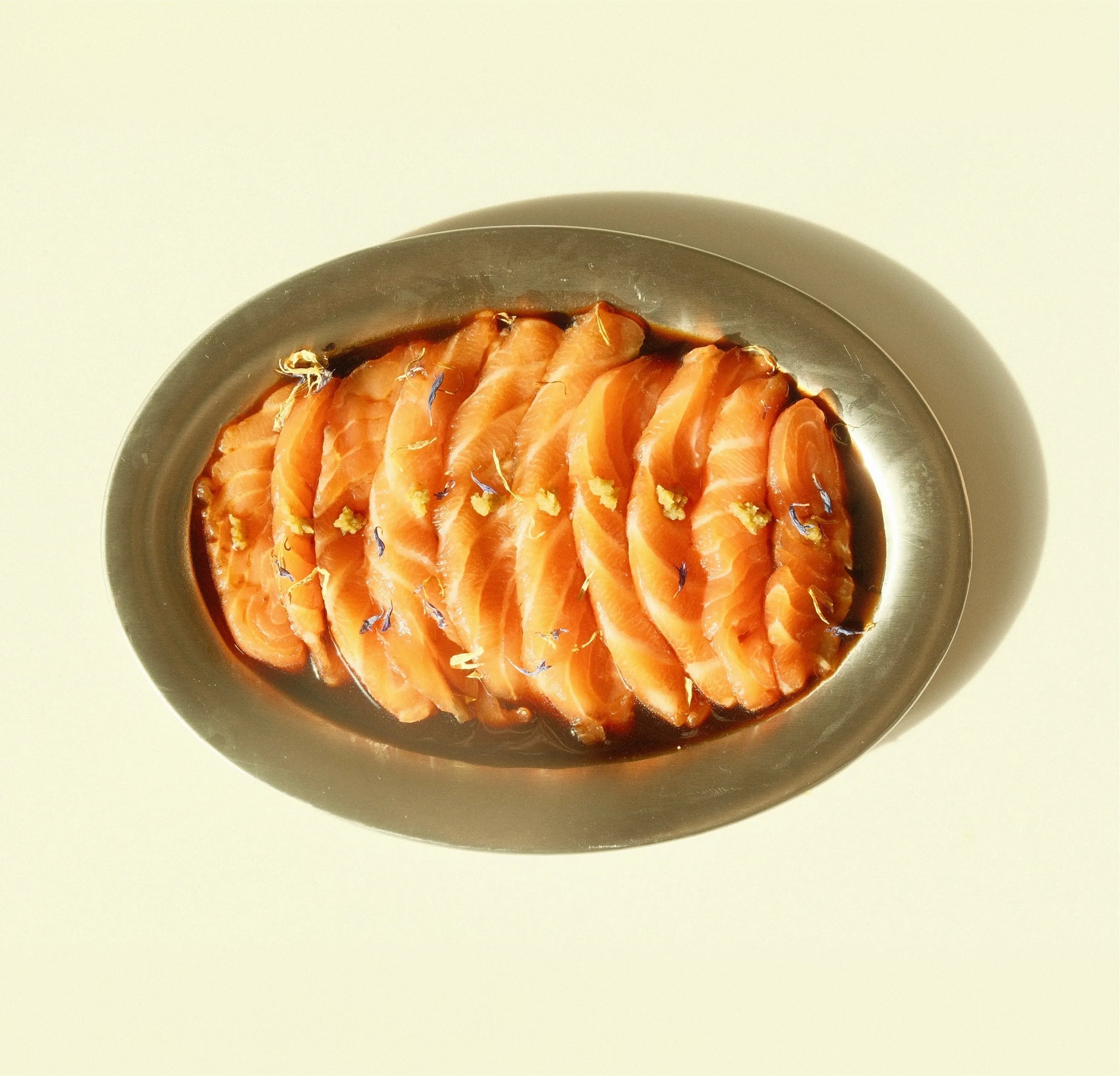 Sliced kombu-cured salmon arranged on an oval metal platter with ponzu sauce and yuzu kosho garnish on a pale background