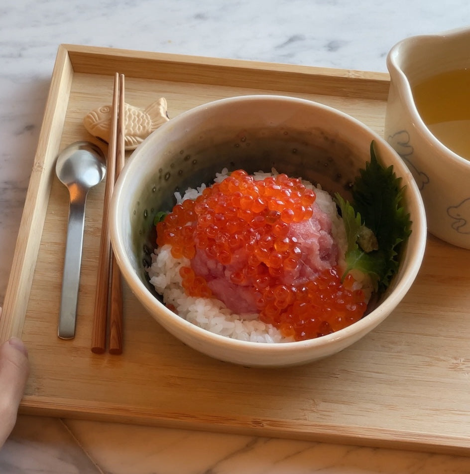 Kaisen donburi topped with minced tuna and bright salmon roe on rice in a ceramic bowl on a wooden tray with chopsticks and a cup of tea