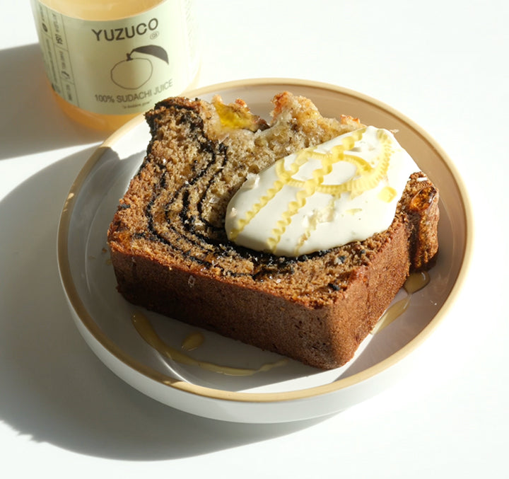 Slice of black sesame banana bread topped with sudachi crème fraîche and citrus zest, with a YUZUCO sudachi juice jar behind