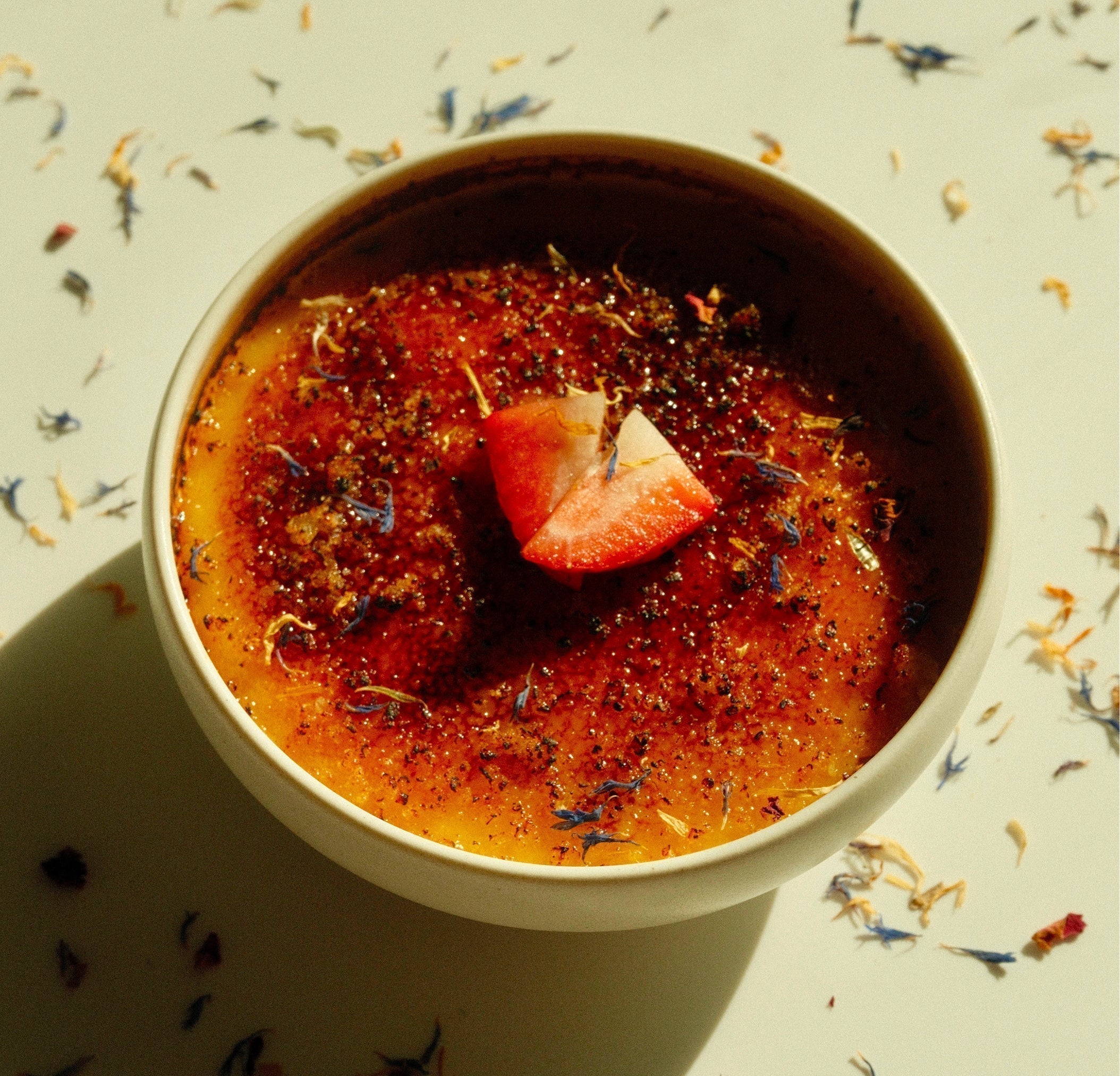 Top view of burnt yuzu crème brûlée with caramelized sugar crust, a sliced strawberry, and scattered edible flower petals on white background