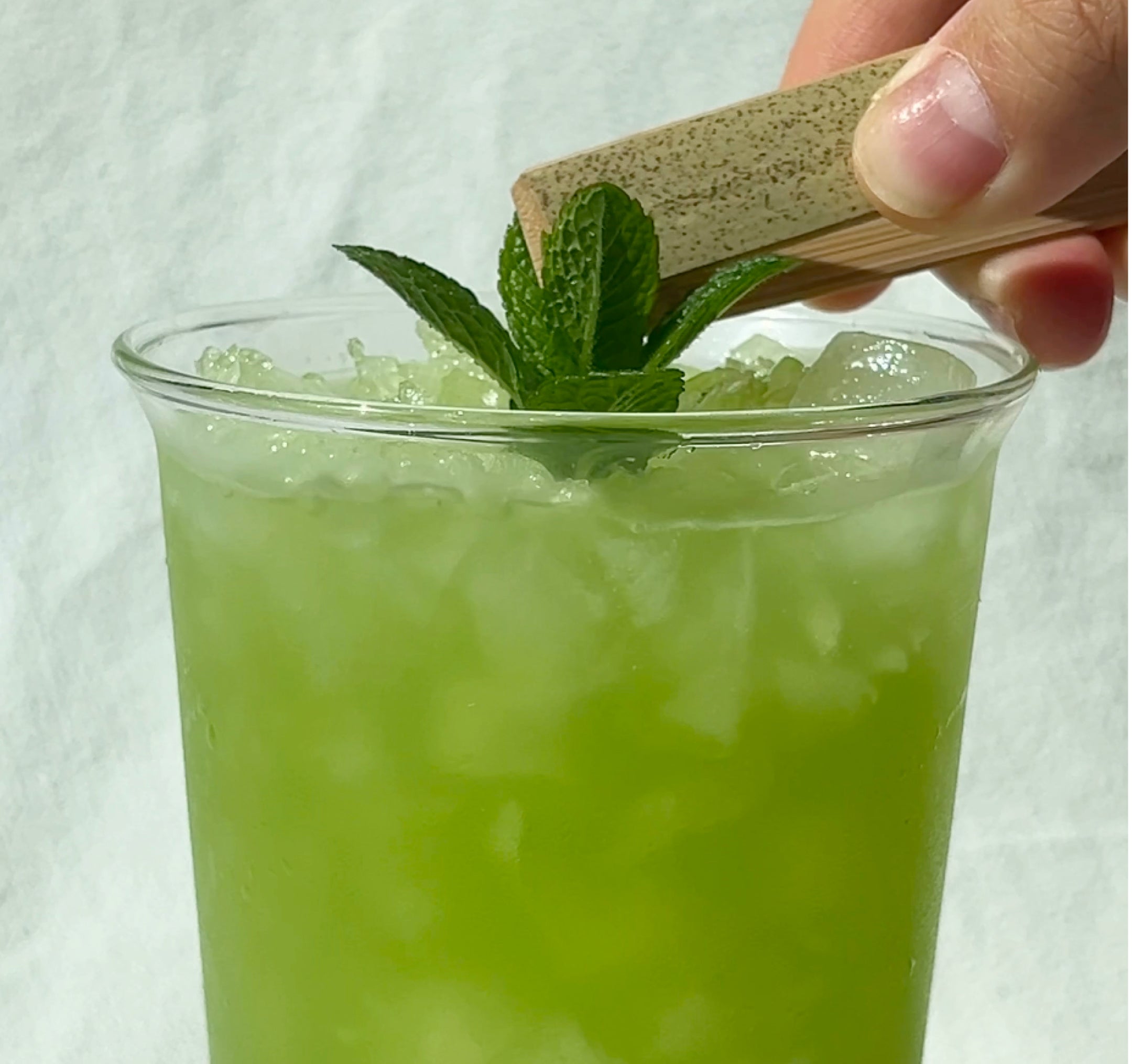 Glass of pale green cucumber-lime drink with crushed ice and a mint sprig, a hand pressing a wooden muddler against the mint