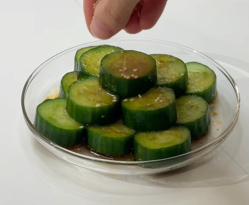 Hand sprinkling sesame seeds over yuzu kosho cucumber rounds on a clear glass plate