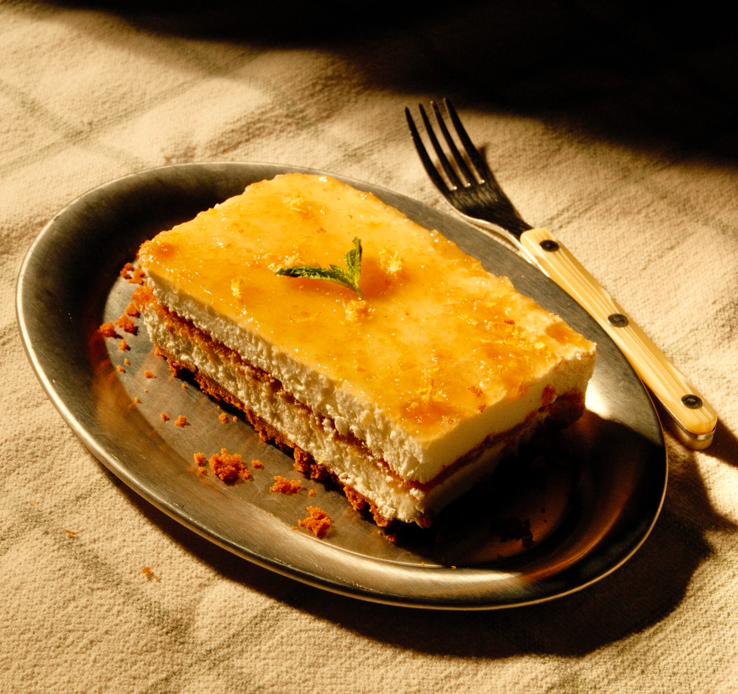 Slice of yuzu float on a metal plate with glossy yuzu glaze, graham cracker crust layers and a mint sprig, fork on beige linen