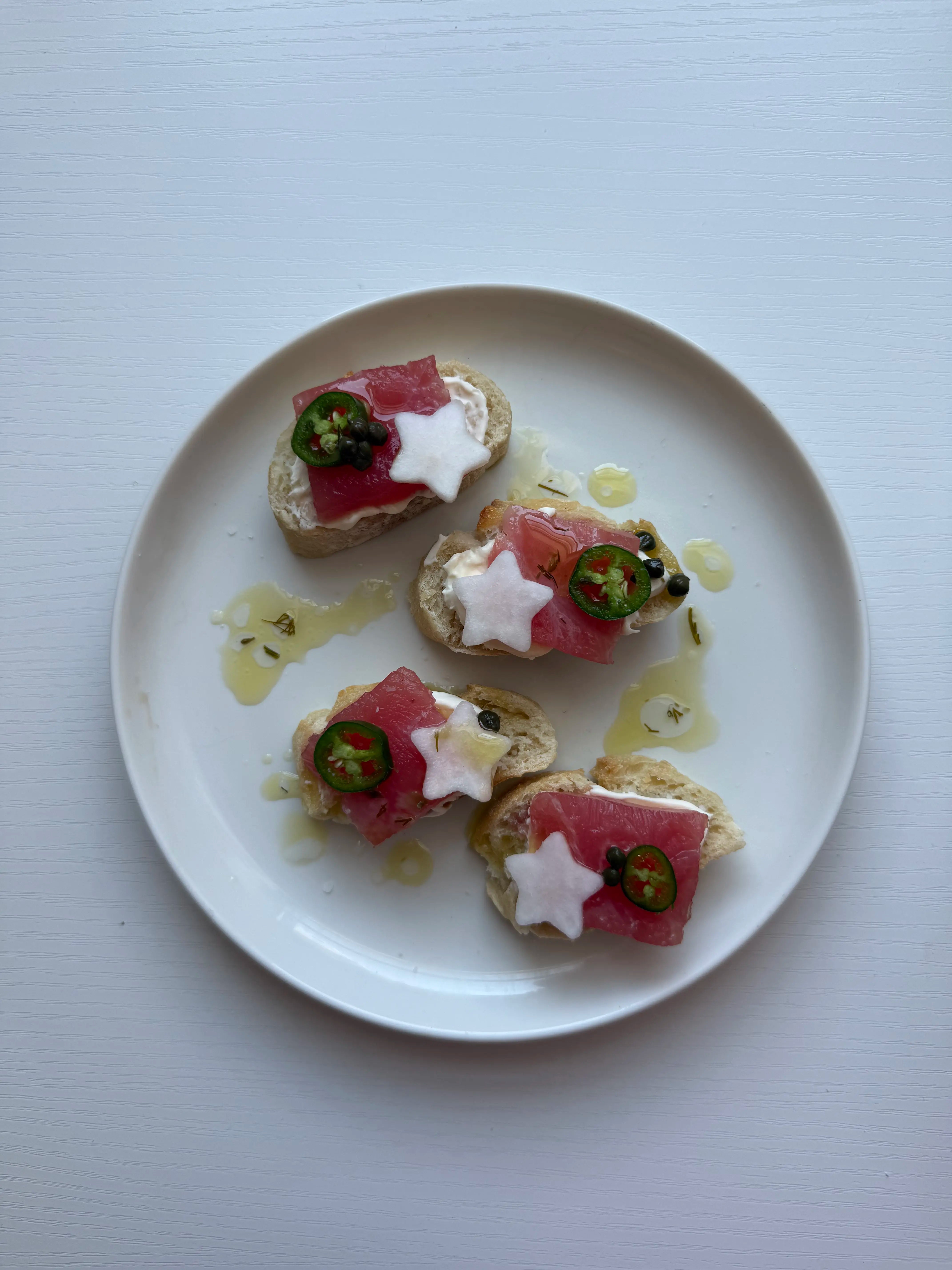 Four yuzu tuna crostini on a white plate topped with crème fraîche, star-shaped radish, serrano slices, capers and a citrus oil drizzle